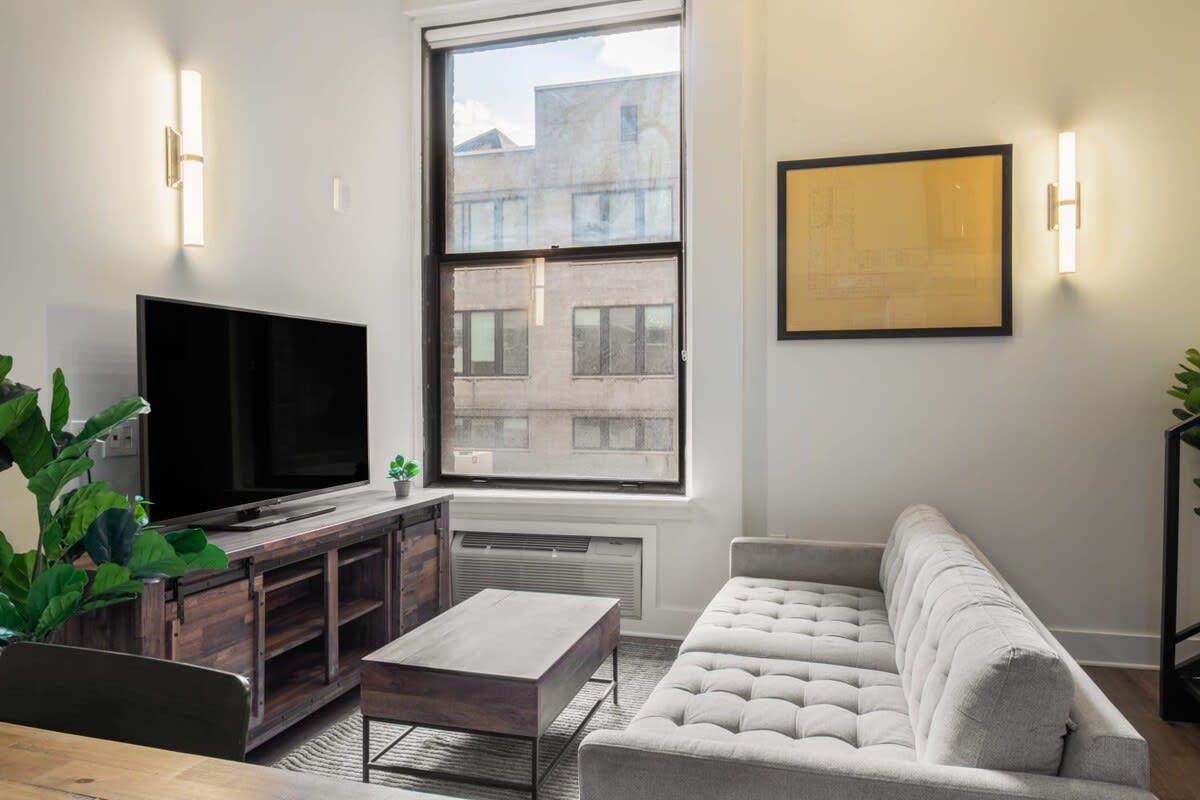 Bright living room featuring a light gray tufted sofa, a dark wood coffee table, and a large flat-screen TV on a rustic console, with a tall window offering a view of adjacent city buildings.