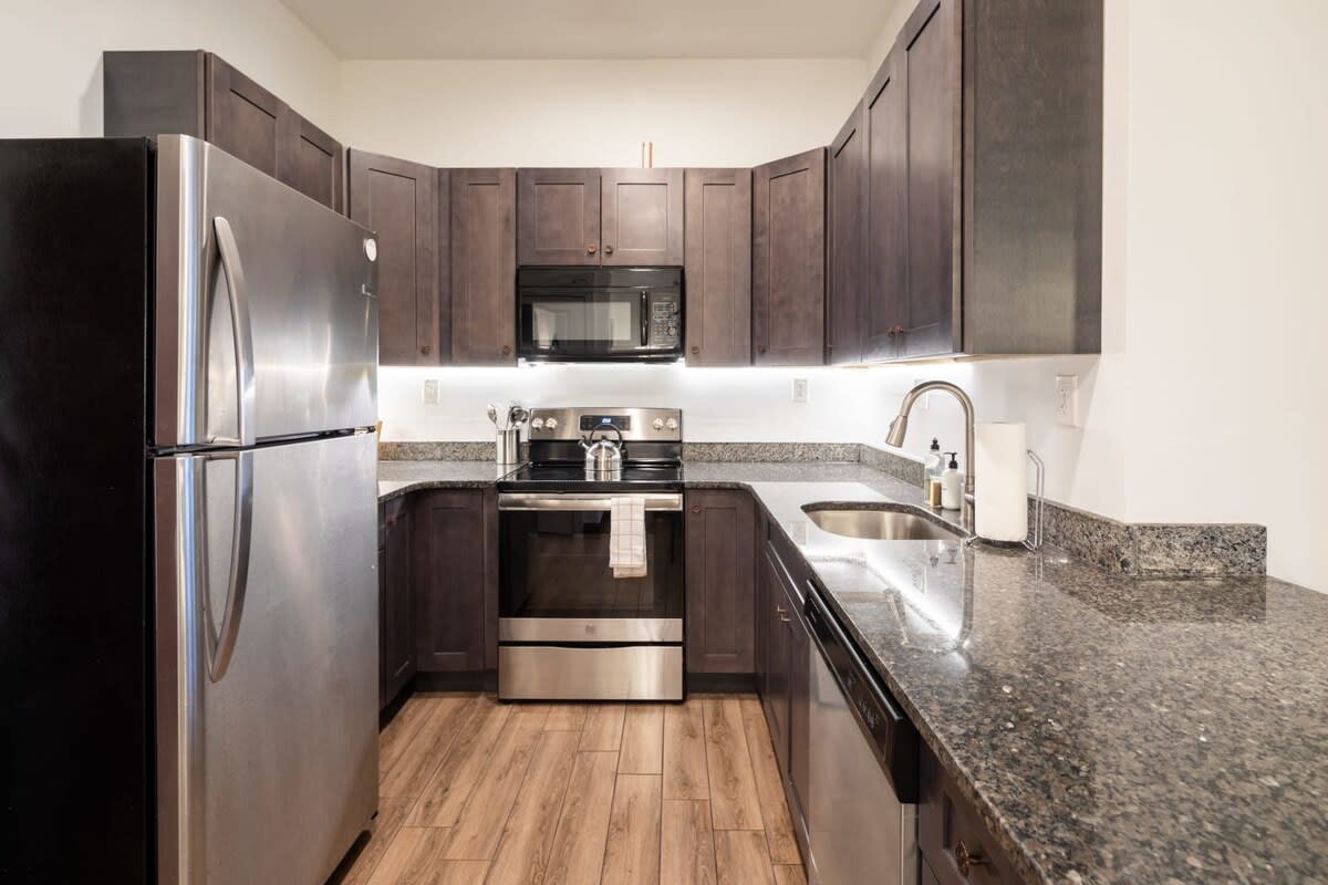 A U-shaped kitchen featuring dark wood cabinetry, granite countertops, and stainless steel appliances including a refrigerator, stove, microwave, and dishwasher. Under-cabinet lighting illuminates the workspace.