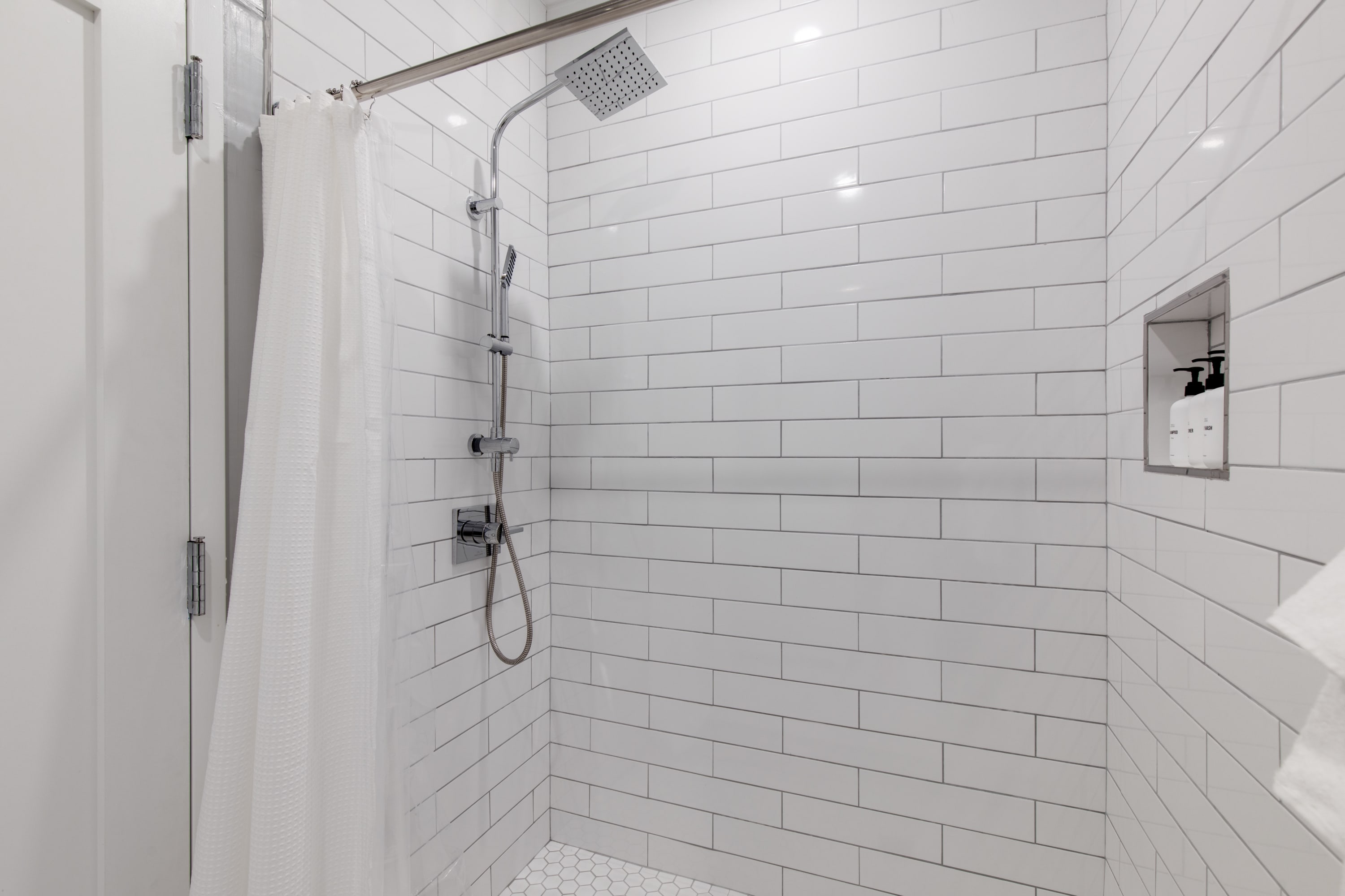 Modern shower stall with white subway tiles on the walls and small white hexagonal tiles on the floor. It features a rain shower head and a handheld shower attachment, with a white shower curtain.