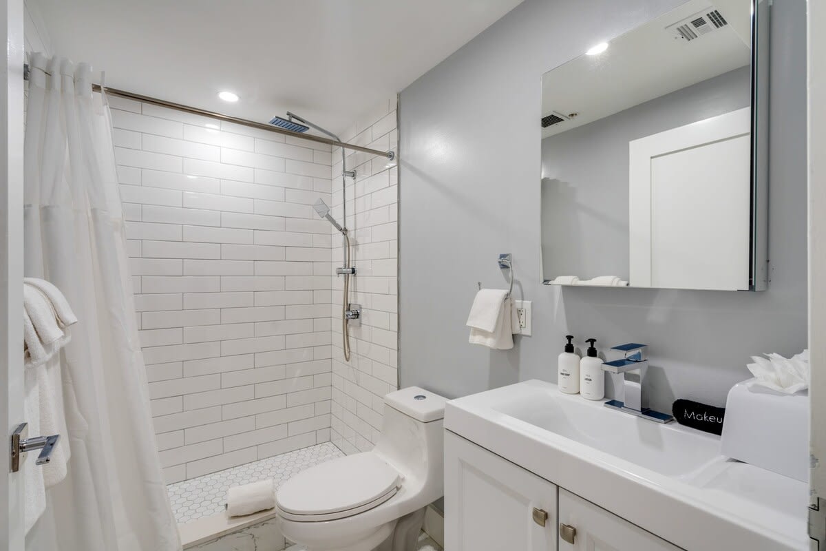 A clean, modern bathroom features a white subway-tiled shower, a white toilet, and a vanity with a rectangular sink and a large mirror. Light gray walls and hexagonal floor tiles complete the contemporary design.