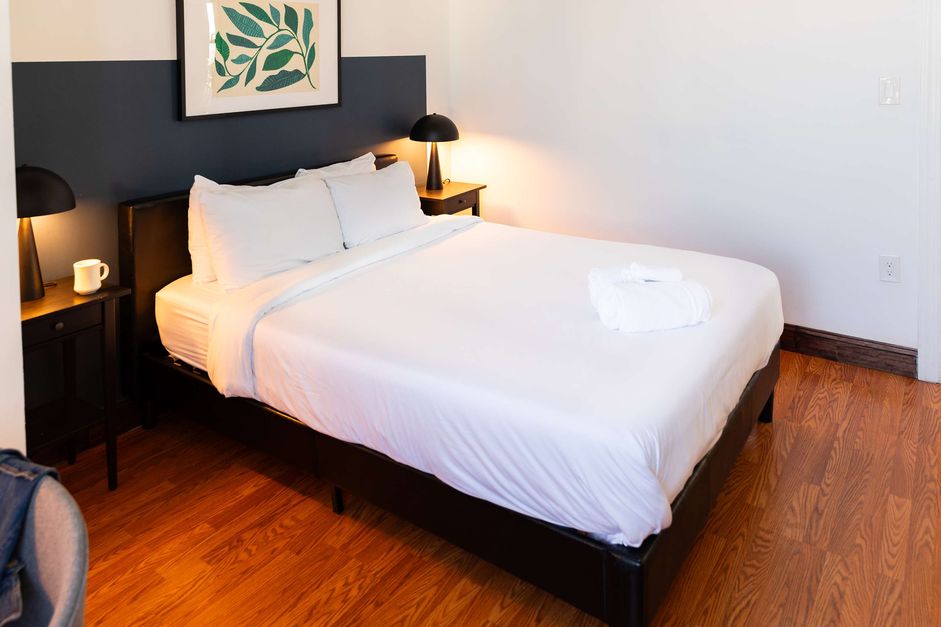 Second bedroom featuring a queen-sized bed with crisp white bedding, flanked by dark nightstands with matching lamps. The room has warm hardwood floors and a striking two-toned wall in white and dark blue/grey, with a framed botanical print above the bed.