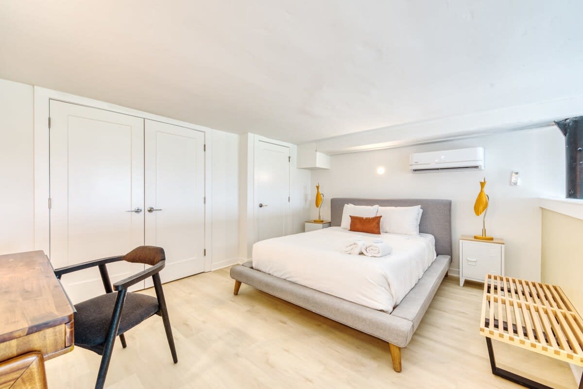 Cozy bedroom featuring a queen-sized bed with a gray upholstered headboard, crisp white linens, and an orange accent pillow. Two white nightstands with distinctive yellow bird lamps flank the bed, while a wooden slatted bench sits at the foot, all on light wood flooring.