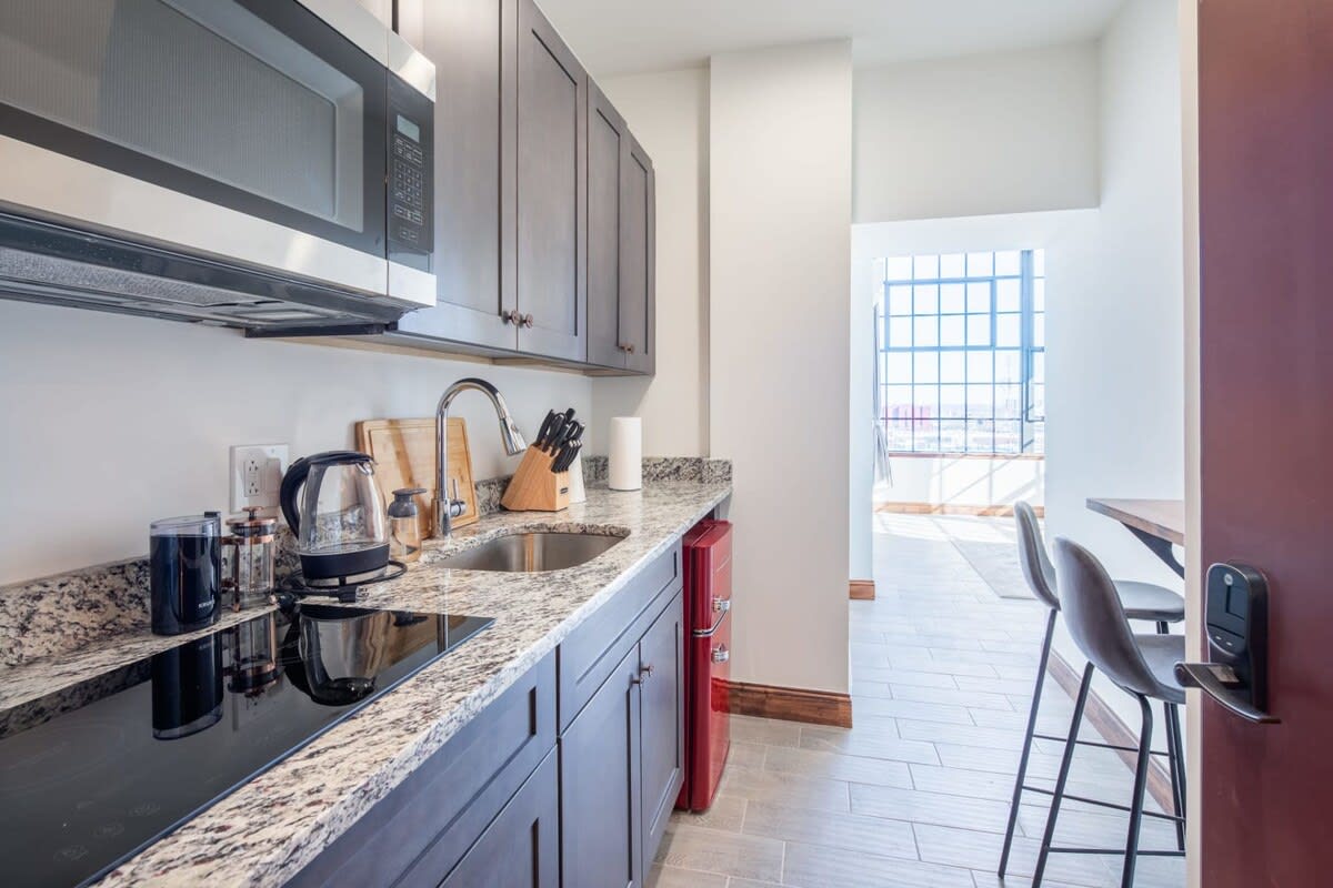 Compact kitchen area with dark wood cabinetry, speckled granite countertops, a stainless steel microwave, and a black electric stovetop. A retro red mini-fridge adds a pop of color, and a sink with a gooseneck faucet is visible.