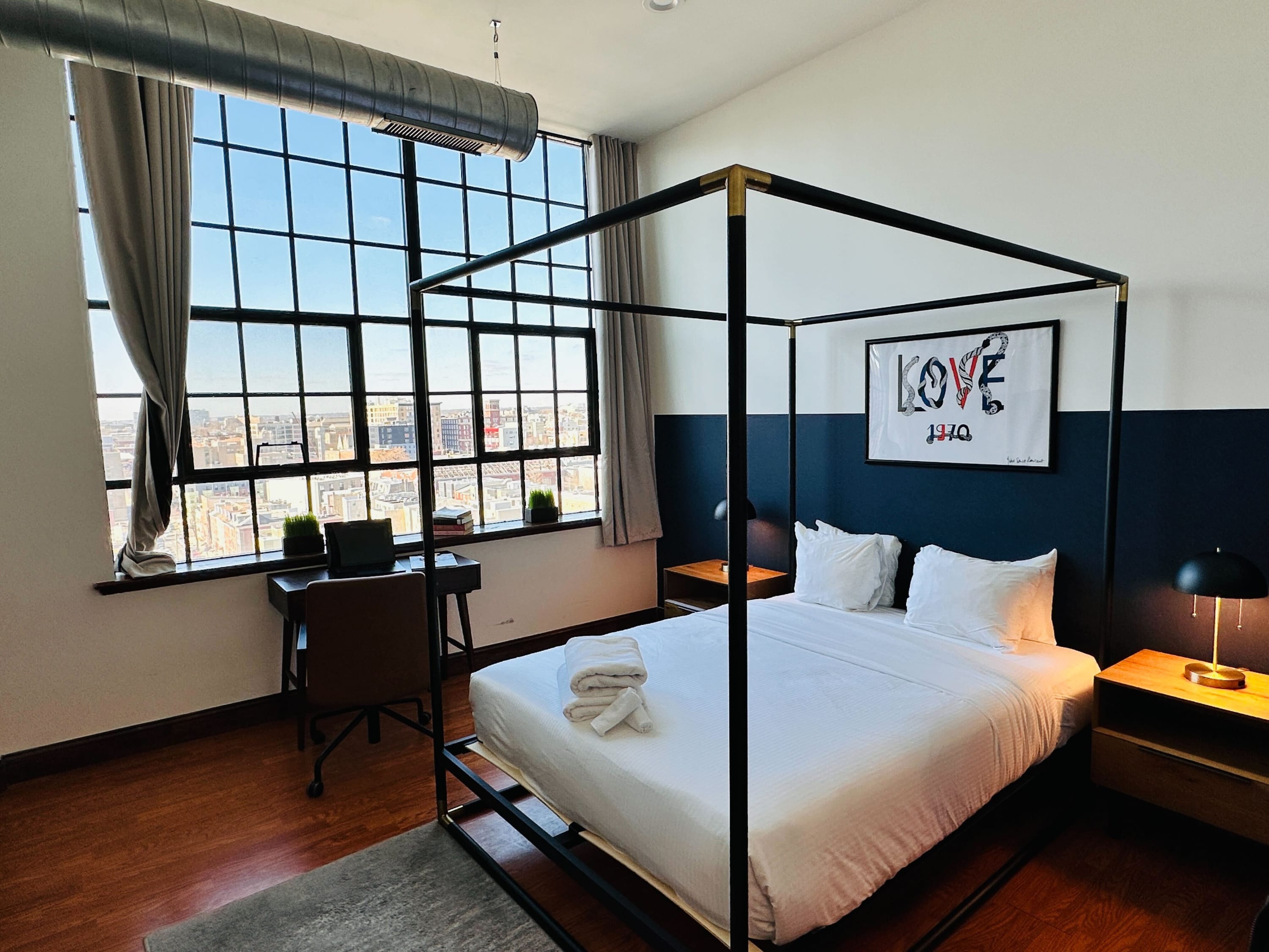 Stylish bedroom featuring a black four-poster bed with white bedding, flanked by wooden nightstands with black lamps. A large window offers city views, and a desk with a brown chair is positioned by the window. The wall has a two-tone paint scheme with white and dark blue.
