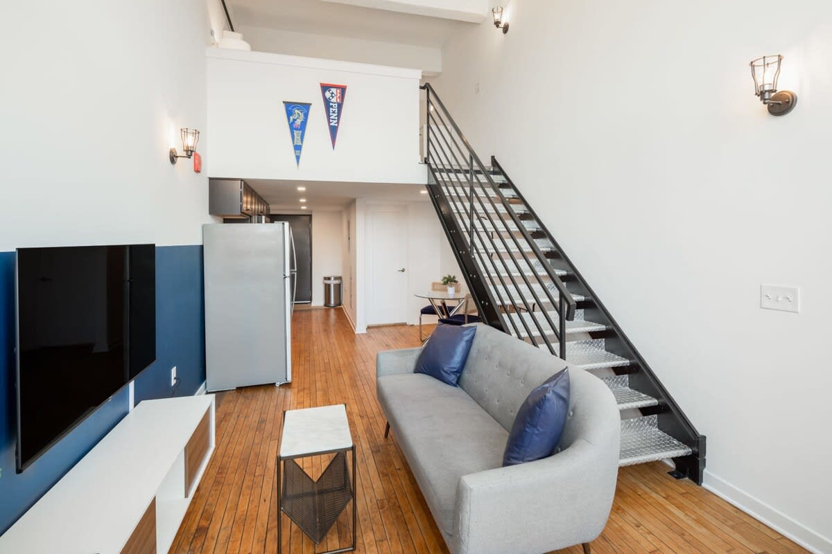 A bright living room featuring a light gray sofa with blue throw pillows, a wall-mounted television above a white and wood media console, and a black metal staircase leading to an upper level.
