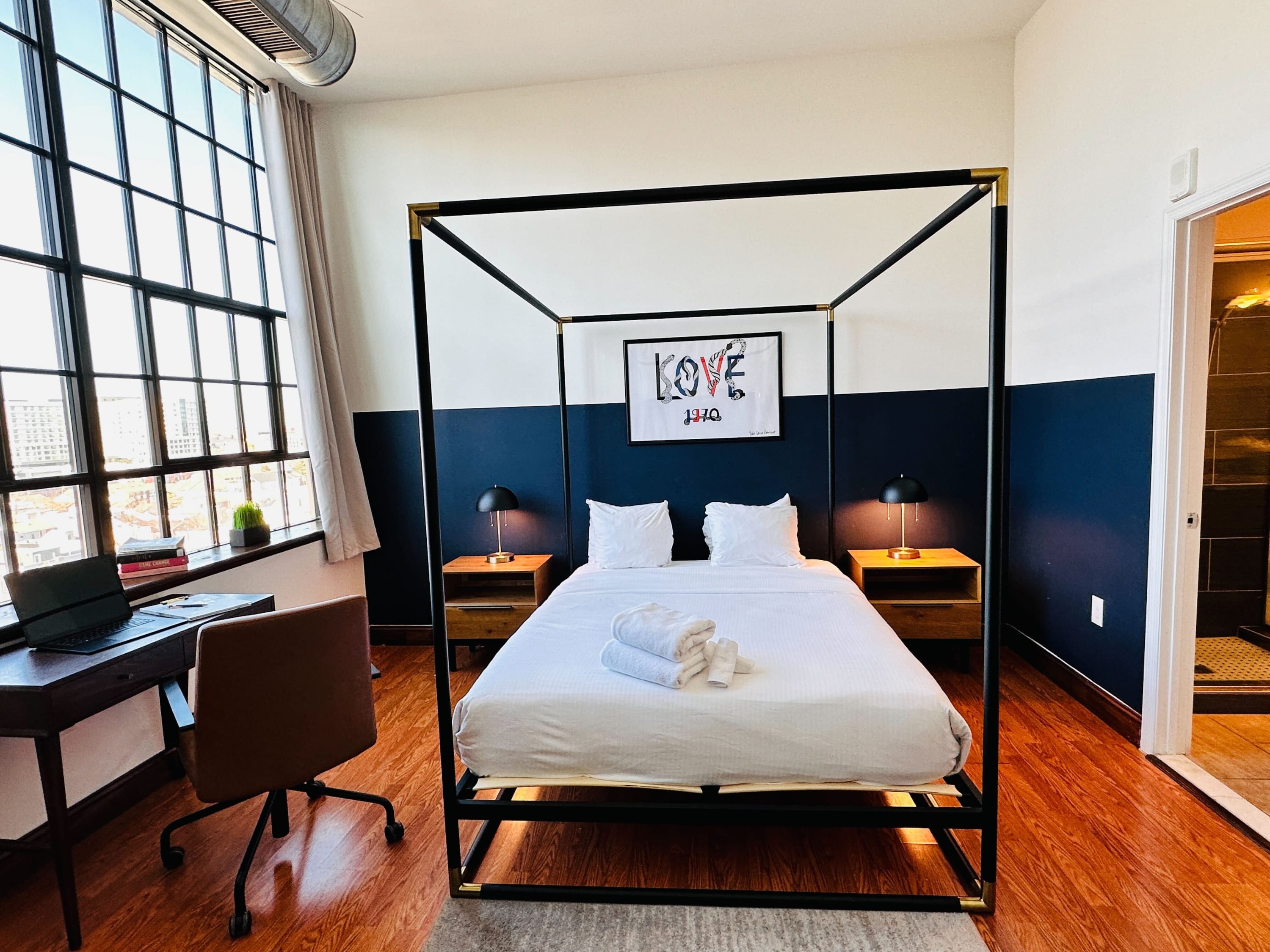 Bright bedroom with a black four-poster bed, white bedding, and two wooden nightstands with black lamps. A large window provides natural light and city views, and a desk with a brown chair is placed beneath it. A "LOVE 1970" artwork hangs above the bed.