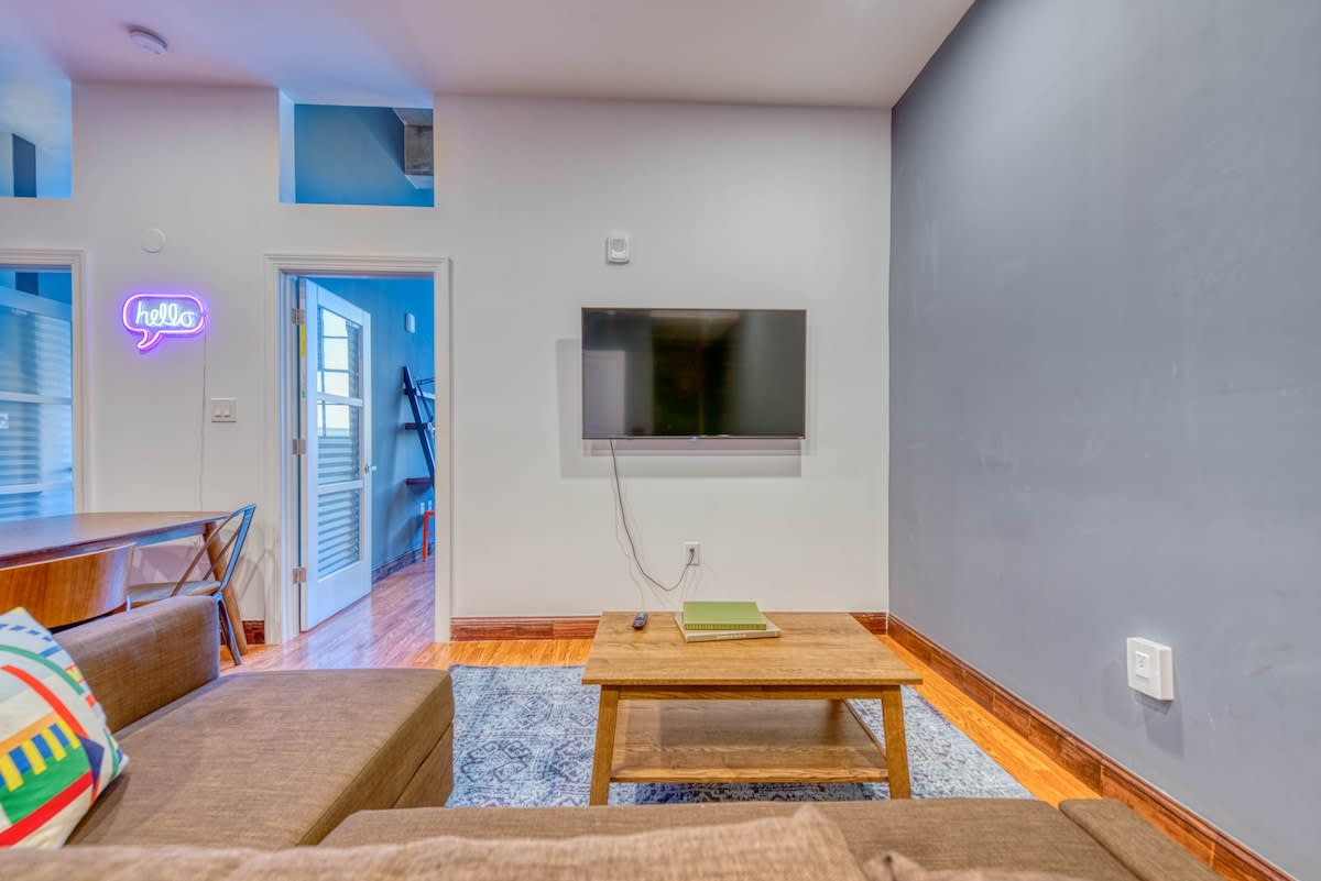 Modern living room featuring a wall-mounted TV above a wooden coffee table, a brown sectional sofa, and a blue patterned area rug. A neon 'hello' sign illuminates a doorway.