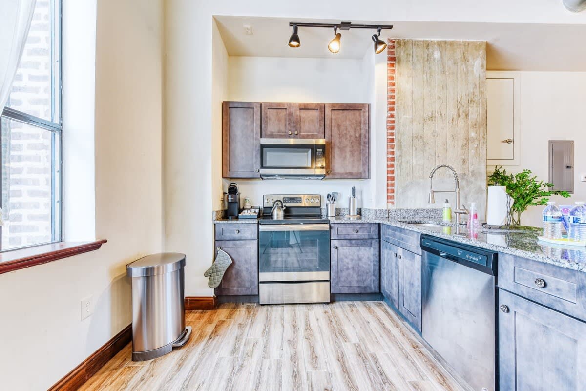Modern kitchen with dark wood cabinets, granite countertops, and stainless steel appliances including a microwave, oven, and dishwasher. A large window provides natural light, and the floor is light wood-look laminate.