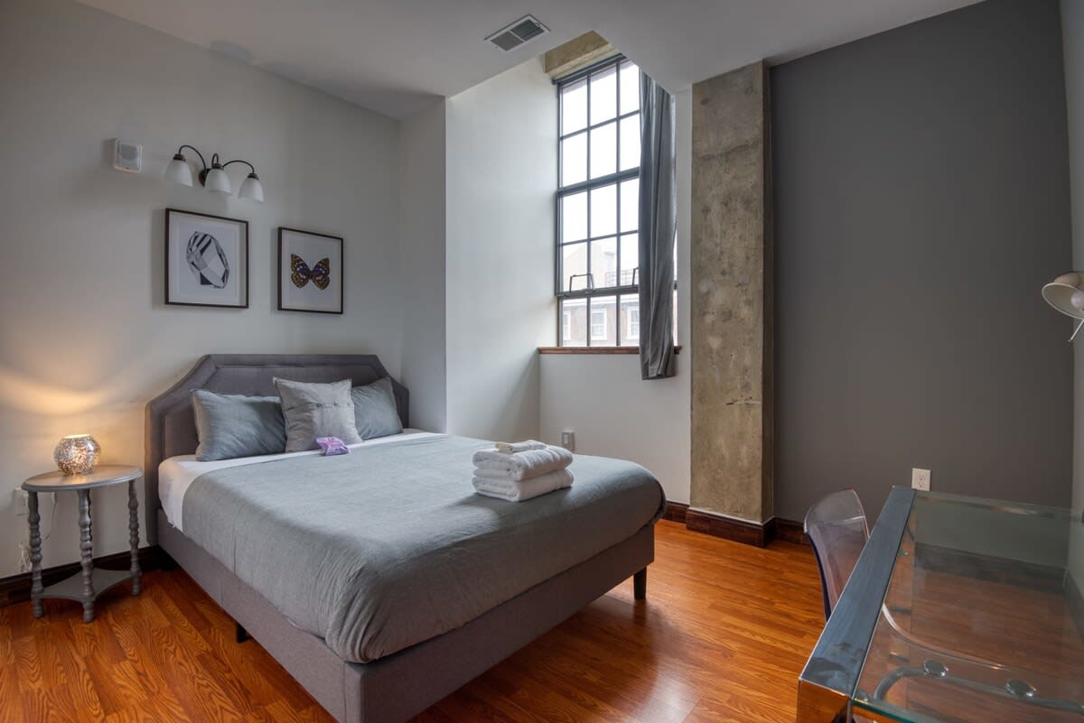This bedroom features a queen-sized bed with a gray upholstered headboard and gray bedding, accompanied by a small round nightstand with a lamp. A tall, narrow industrial-style window with a gray curtain is visible on the right, and two framed artworks hang above the bed, all on light hardwood floors.