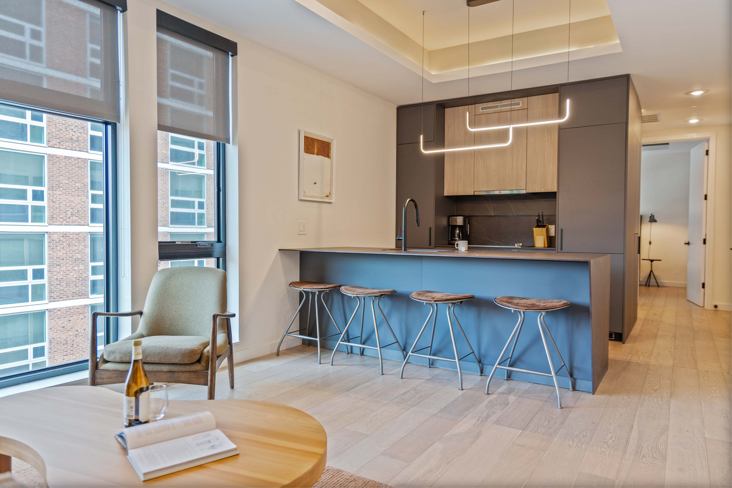 Modern open-concept kitchen with dark gray and light wood cabinetry, a large island with a sink and four bar stools, and sleek linear pendant lighting. Large windows provide ample natural light and city views, connecting to a living area.