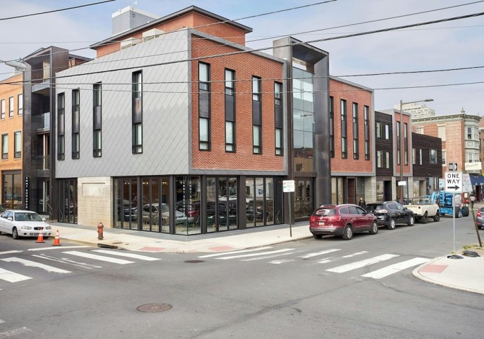 The modern exterior of a multi-story building at a street corner, featuring a mix of red brick and gray metal paneling, large windows, and ground-level retail spaces. Cars are parked along the street with a fire hydrant visible.