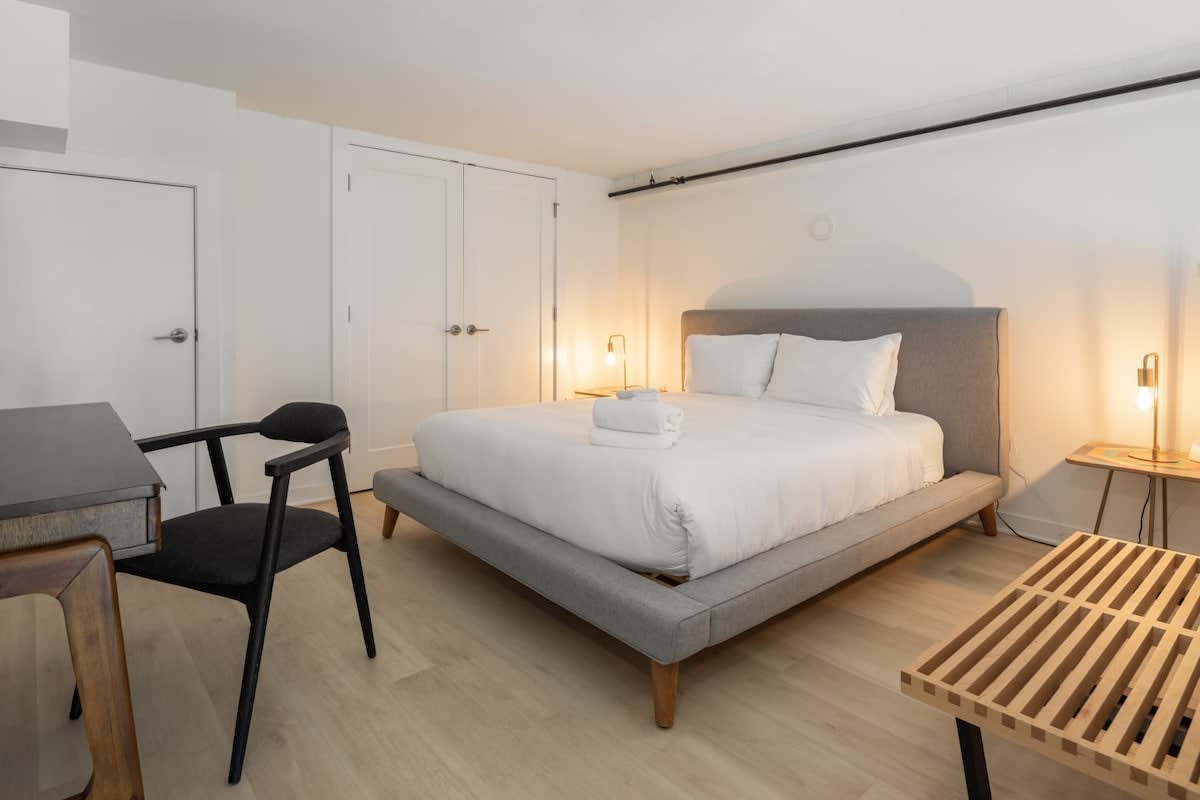 A bright bedroom featuring a grey upholstered queen bed with white bedding and two nightstands with lamps. The room has light wood-look flooring and white walls with double closet doors.