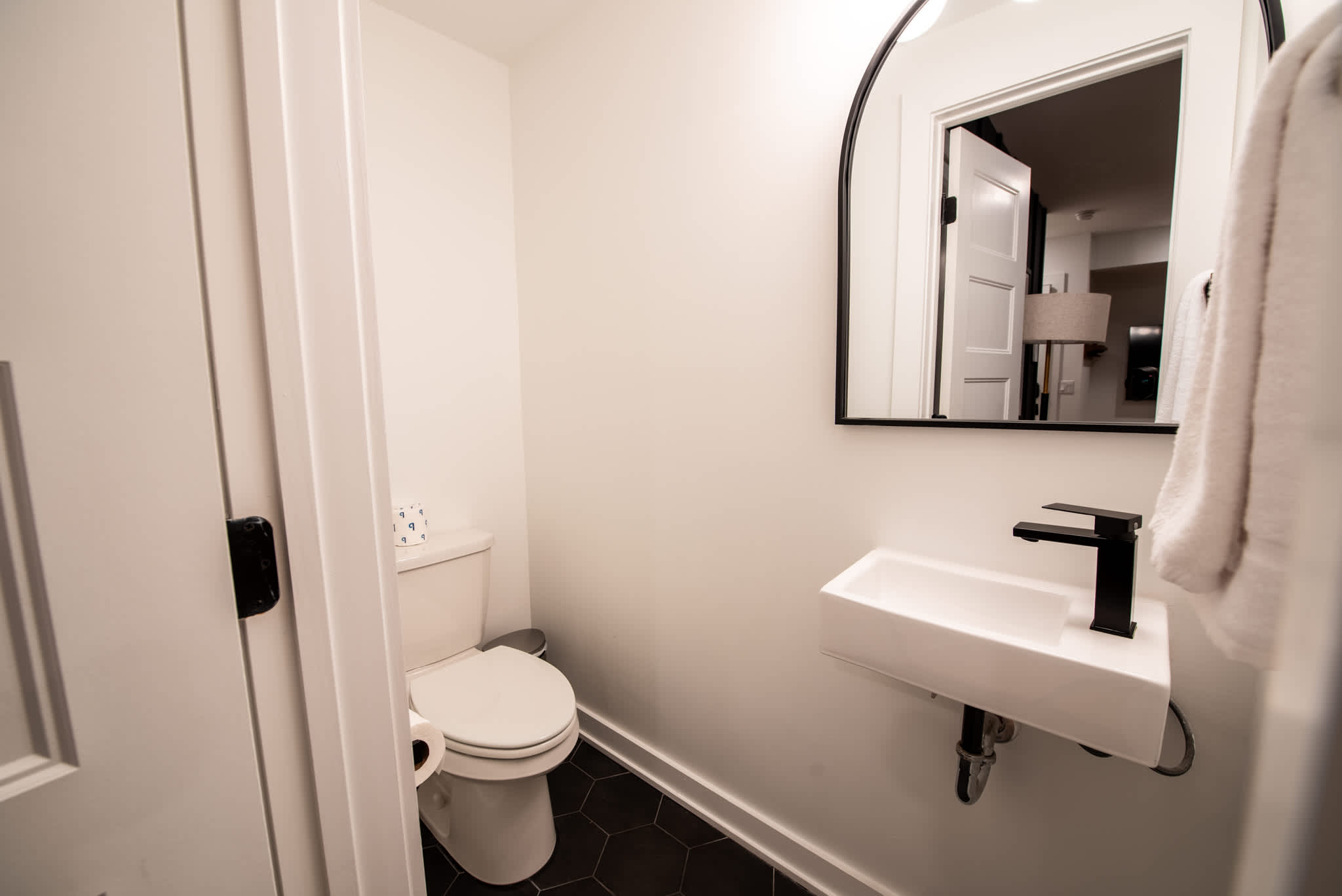 A sleek half-bathroom with a wall-mounted rectangular sink, a black faucet, a toilet, and a modern arched mirror, set against white walls and dark hexagonal floor tiles.