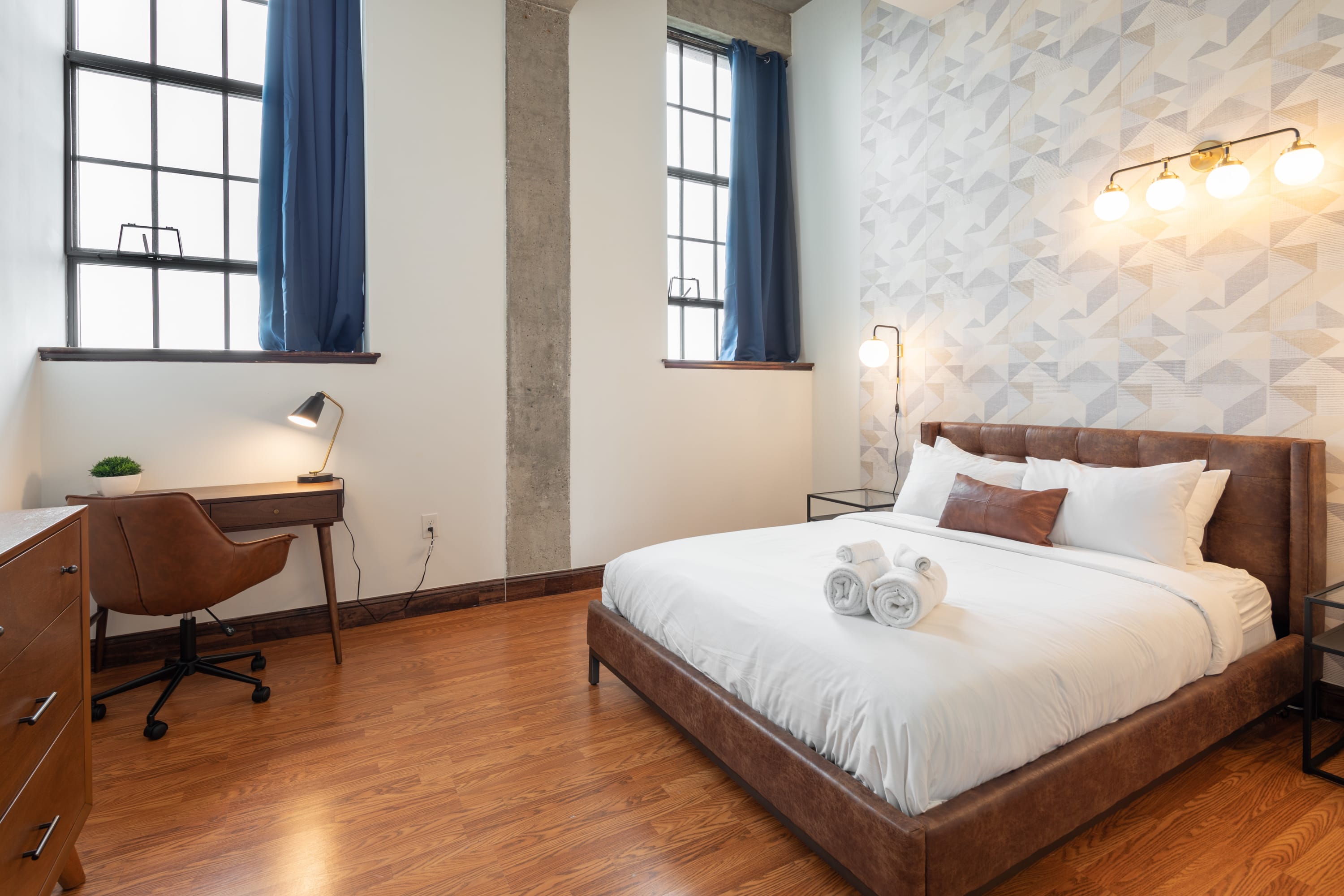 Modern bedroom with a brown leather bed frame, white bedding, and a geometric patterned accent wall. Two large industrial-style windows with blue curtains provide natural light, and a desk with an office chair is in the corner.