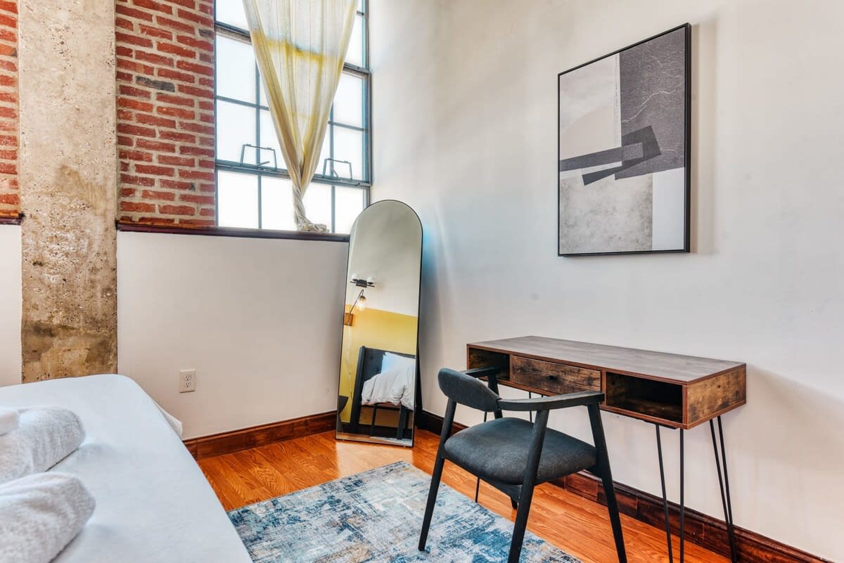 A cozy corner of a bedroom featuring a dark wood desk with hairpin legs and a dark grey upholstered chair, positioned next to a tall arched full-length mirror. The room has light wood flooring, a blue and white abstract rug, and a large industrial window with exposed brick on one side.