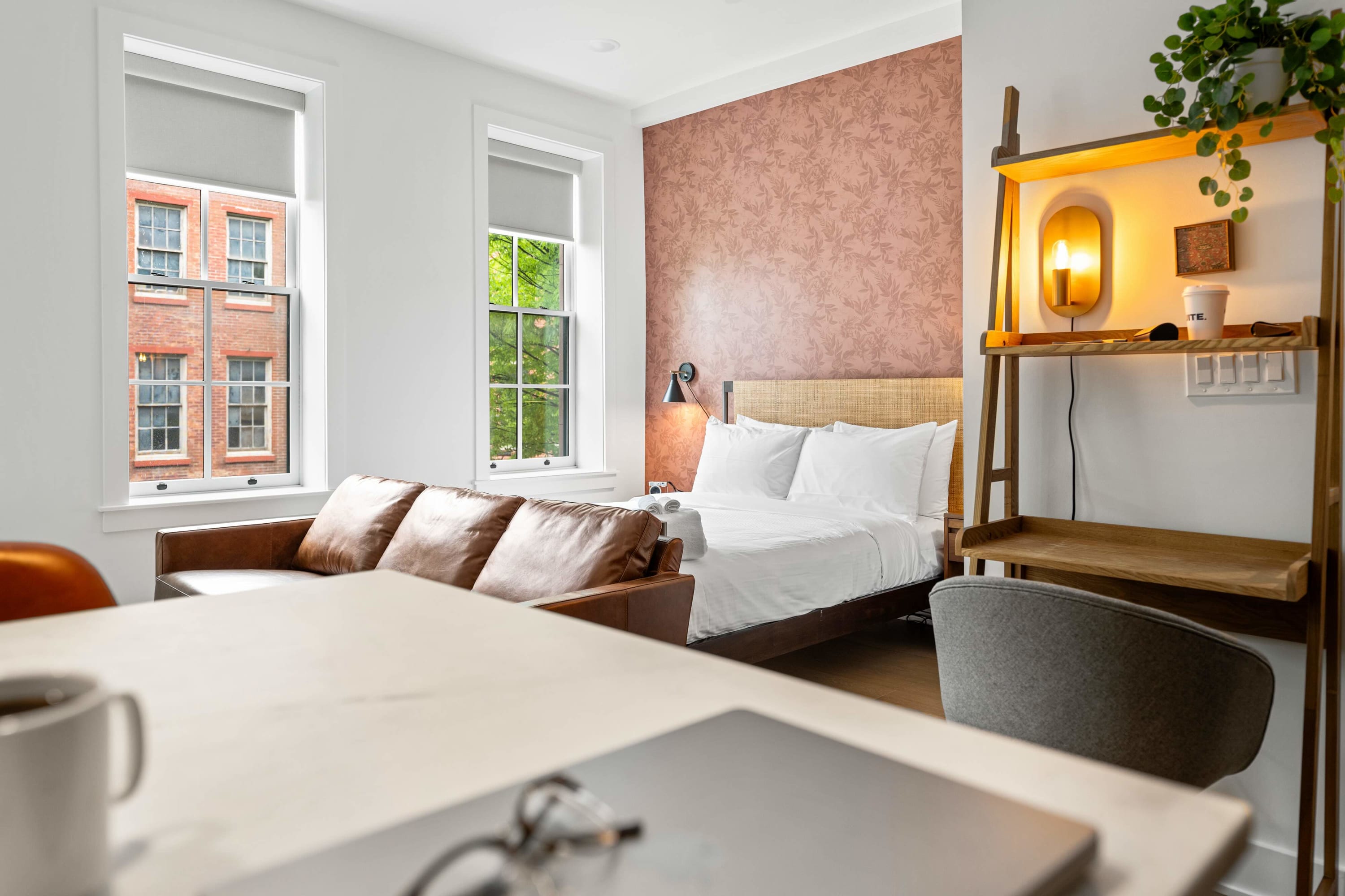 Wide view of the first bedroom, showcasing the queen bed with woven headboard, brown leather sofa, and a wooden ladder desk with a laptop. The room is bright with large windows and a patterned feature wall.