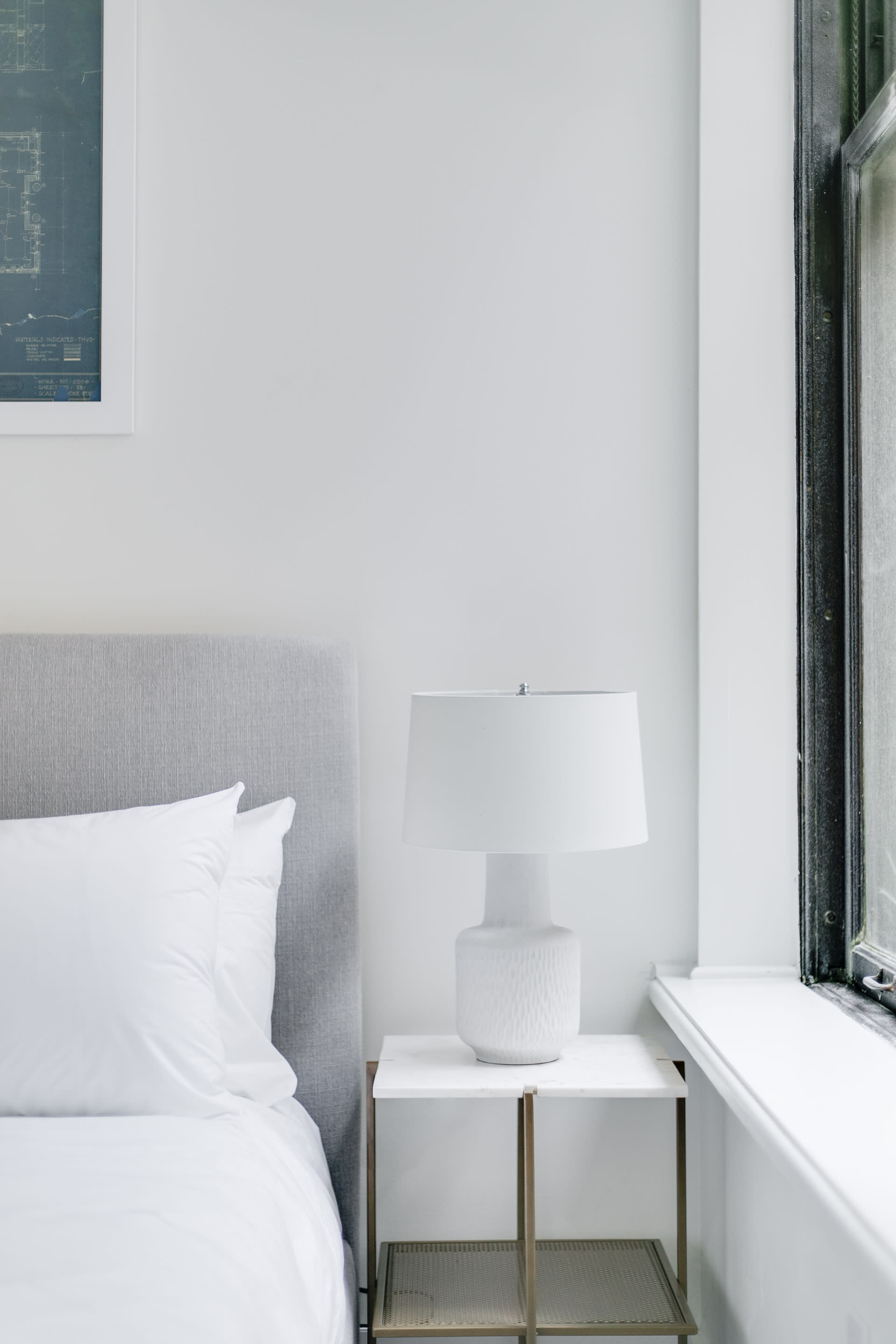 Close-up of a bedside area in a bright bedroom, featuring a white textured table lamp on a minimalist white and gold nightstand next to a gray upholstered bed with white bedding.