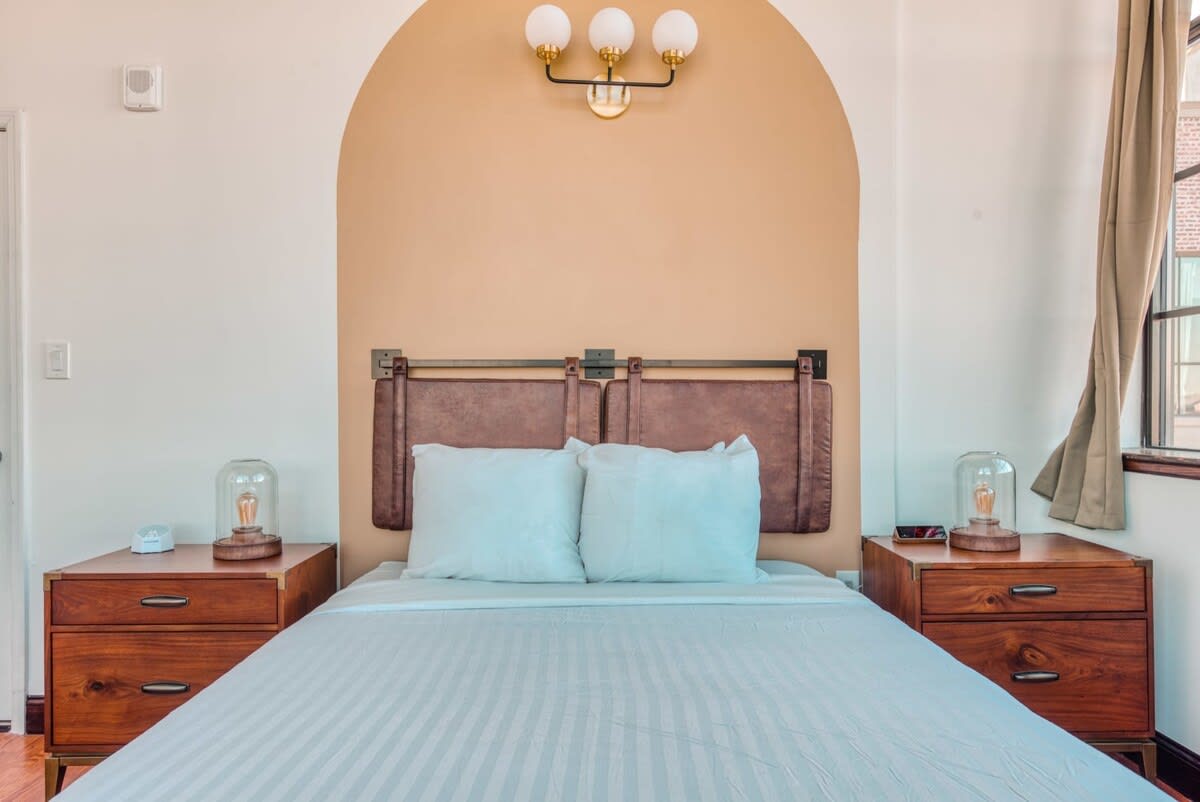 Cozy bedroom with a queen bed, dark wood nightstands, and a unique leather-strap headboard, accented by a warm arched paint detail on the wall and modern wall sconces.