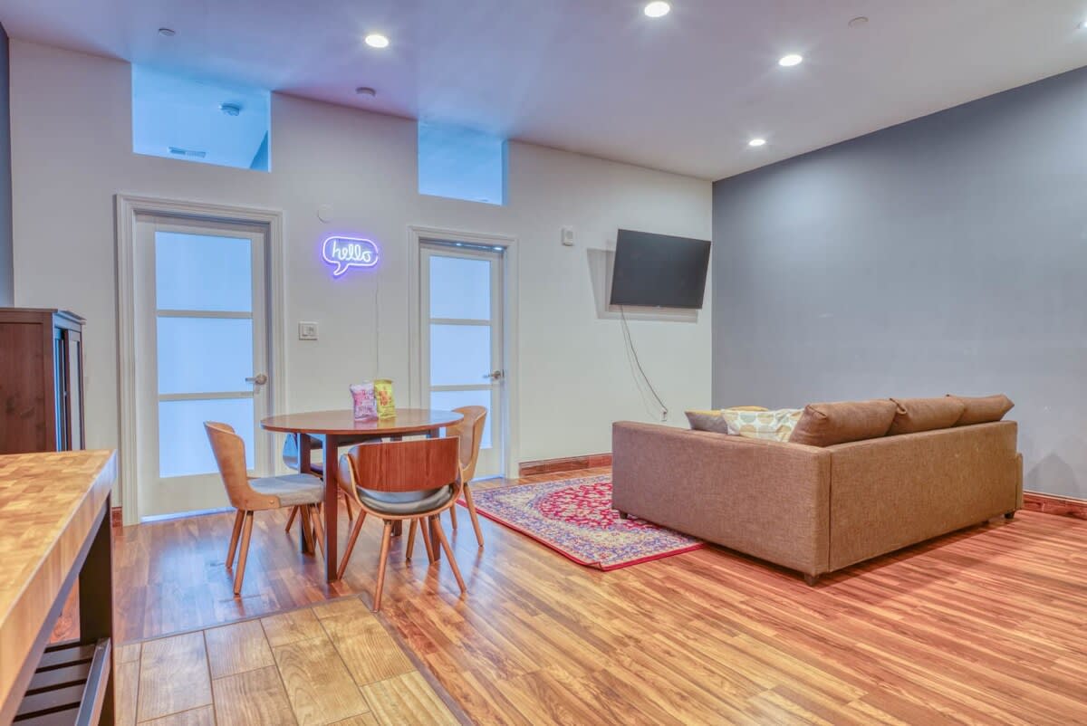 An open-concept living and dining space with a round wooden dining table and chairs, a "hello" neon sign, and a comfortable brown sofa facing a wall-mounted TV, all on warm hardwood floors.