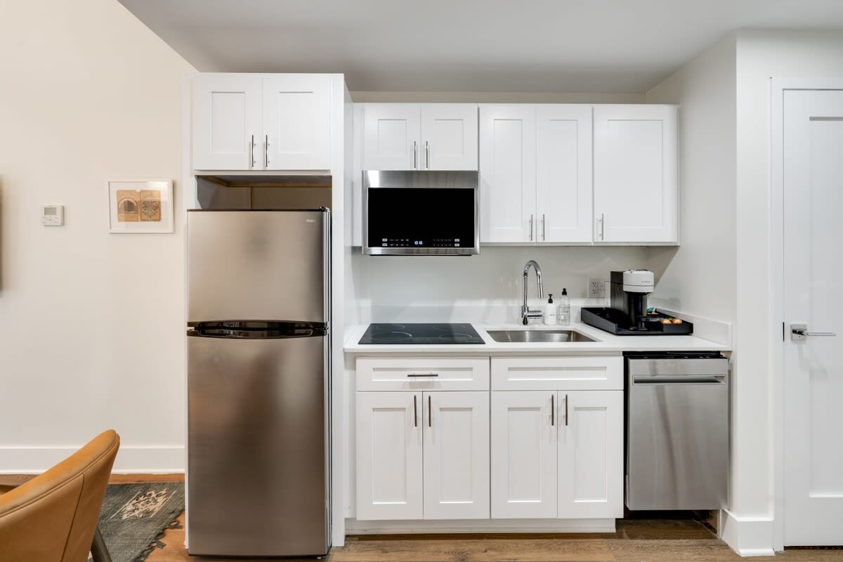A compact, modern kitchen is equipped with white shaker cabinets, stainless steel appliances including a refrigerator, microwave, dishwasher, and a two-burner electric cooktop. A coffee maker and sink are visible on the white countertop.