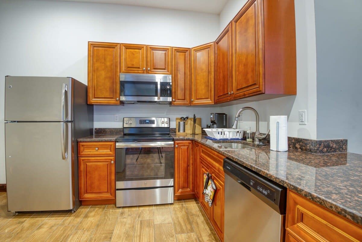 A well-equipped kitchen with warm cherry wood cabinets, dark granite countertops, and stainless steel appliances including a refrigerator, range, microwave, and dishwasher. The floor is covered in light wood-look tile.