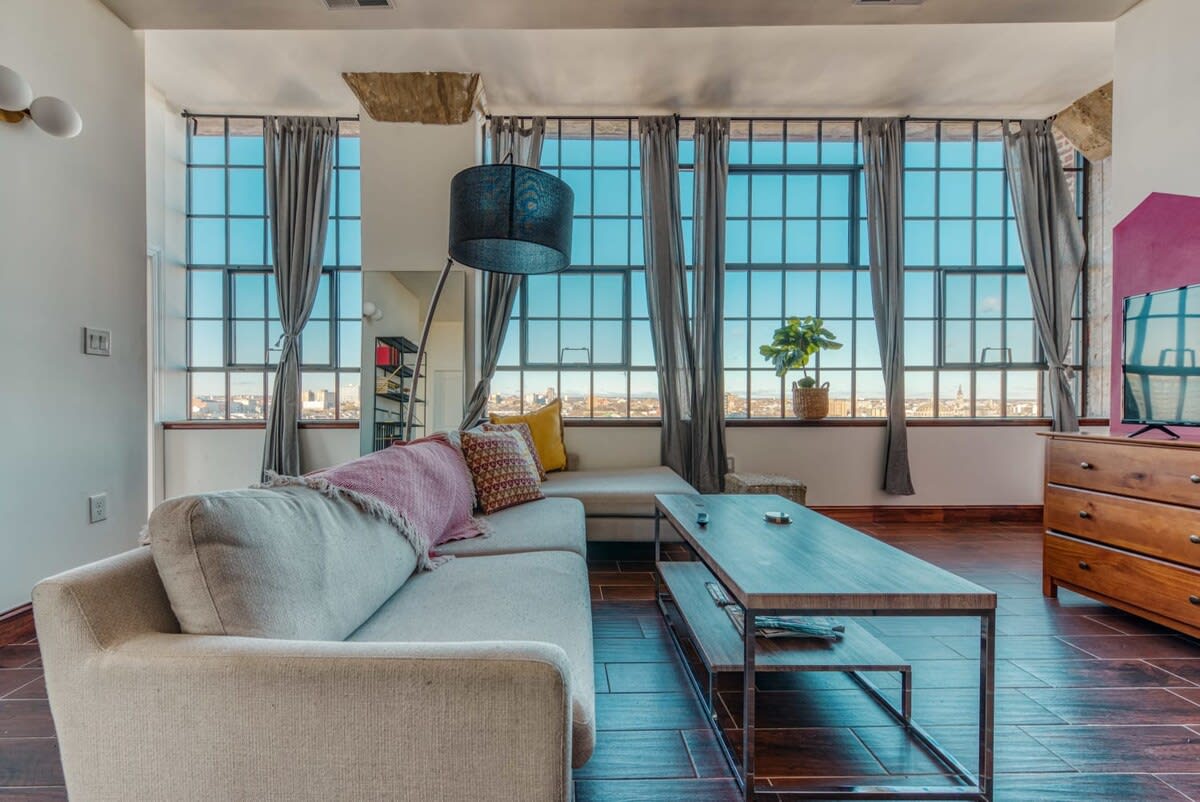 Wide shot of the living room showcasing a light-colored sectional sofa, a rectangular coffee table, and a wall of large grid-patterned windows offering panoramic city views.