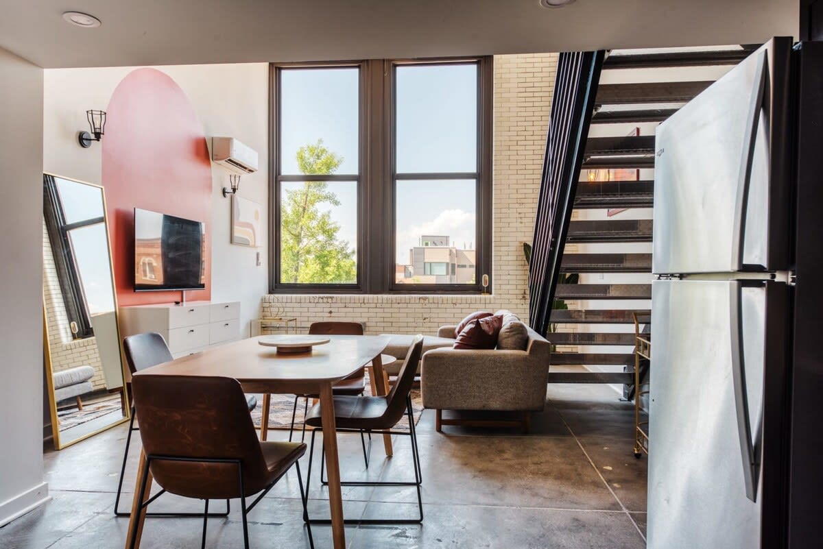Modern dining area with a rectangular wooden table and four brown leather chairs, positioned in front of large industrial-style windows with a white brick wall. A gray sofa and a white media console with a TV are visible in the adjacent living space, with a black metal staircase on the right.