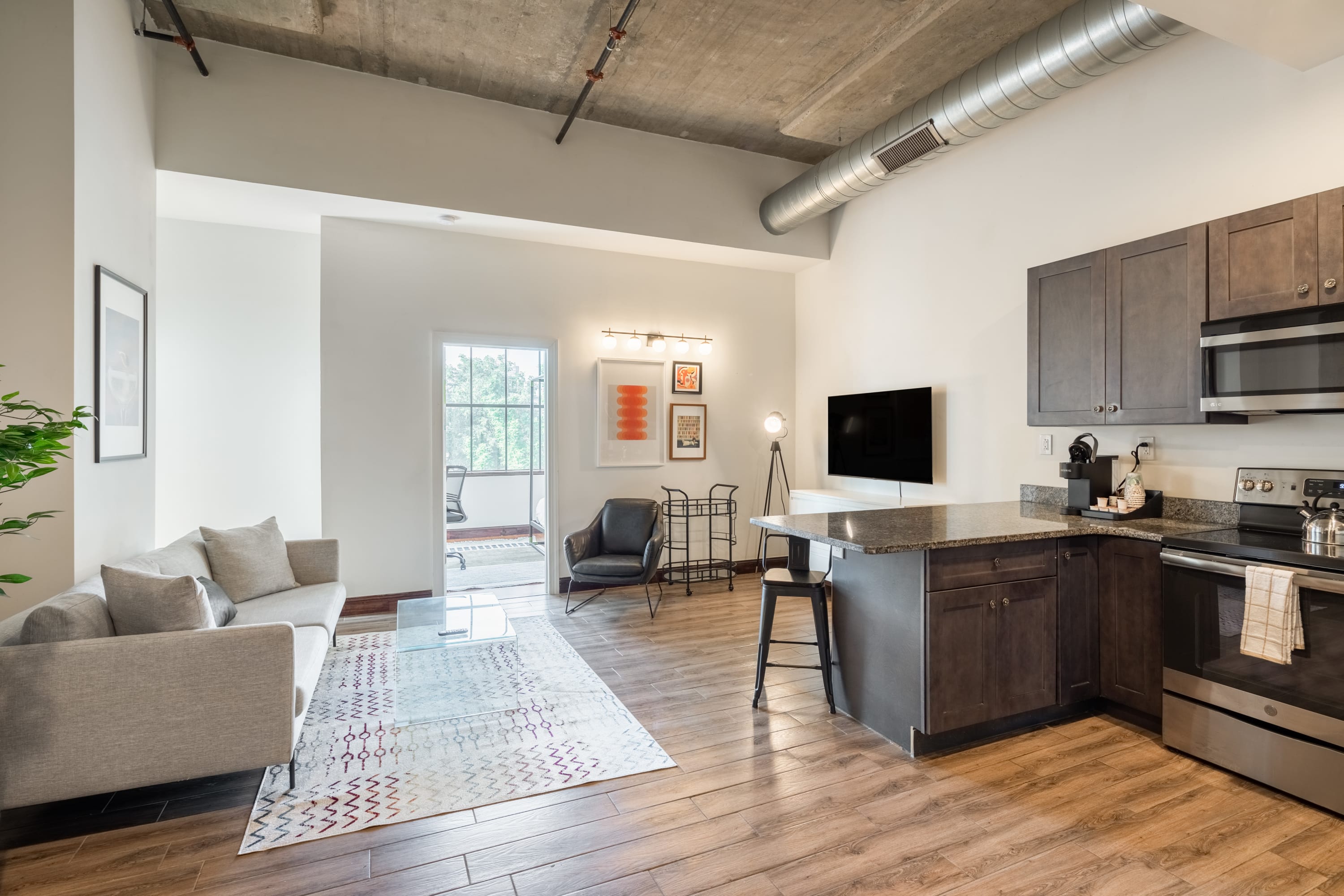 An open-concept living area and kitchen with light wood floors and a concrete ceiling. The living space has a light grey sofa, a glass coffee table, and a view towards a balcony door, while the kitchen features dark grey cabinets and stainless steel appliances.