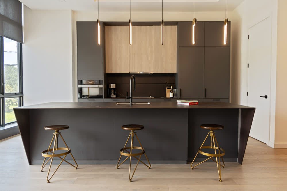 Sleek, modern kitchen featuring a large dark gray island with a waterfall edge and three brass bar stools, dark gray cabinetry, light wood upper cabinets, and stylish pendant lights hanging above.