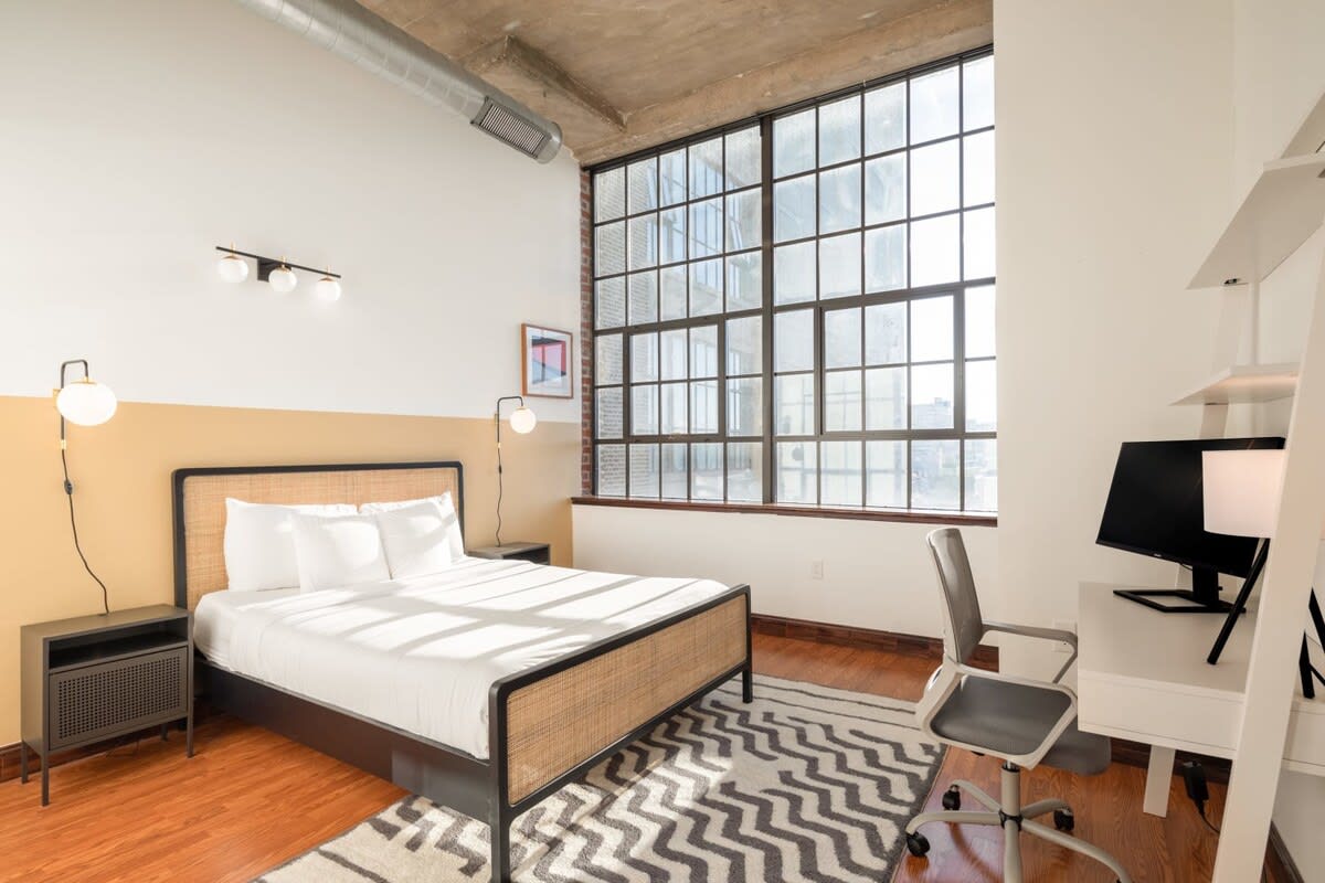 Bright bedroom with a queen-sized bed featuring a woven headboard, a white desk with a computer monitor, and large industrial-style windows. The walls are painted white with a mustard yellow accent stripe.
