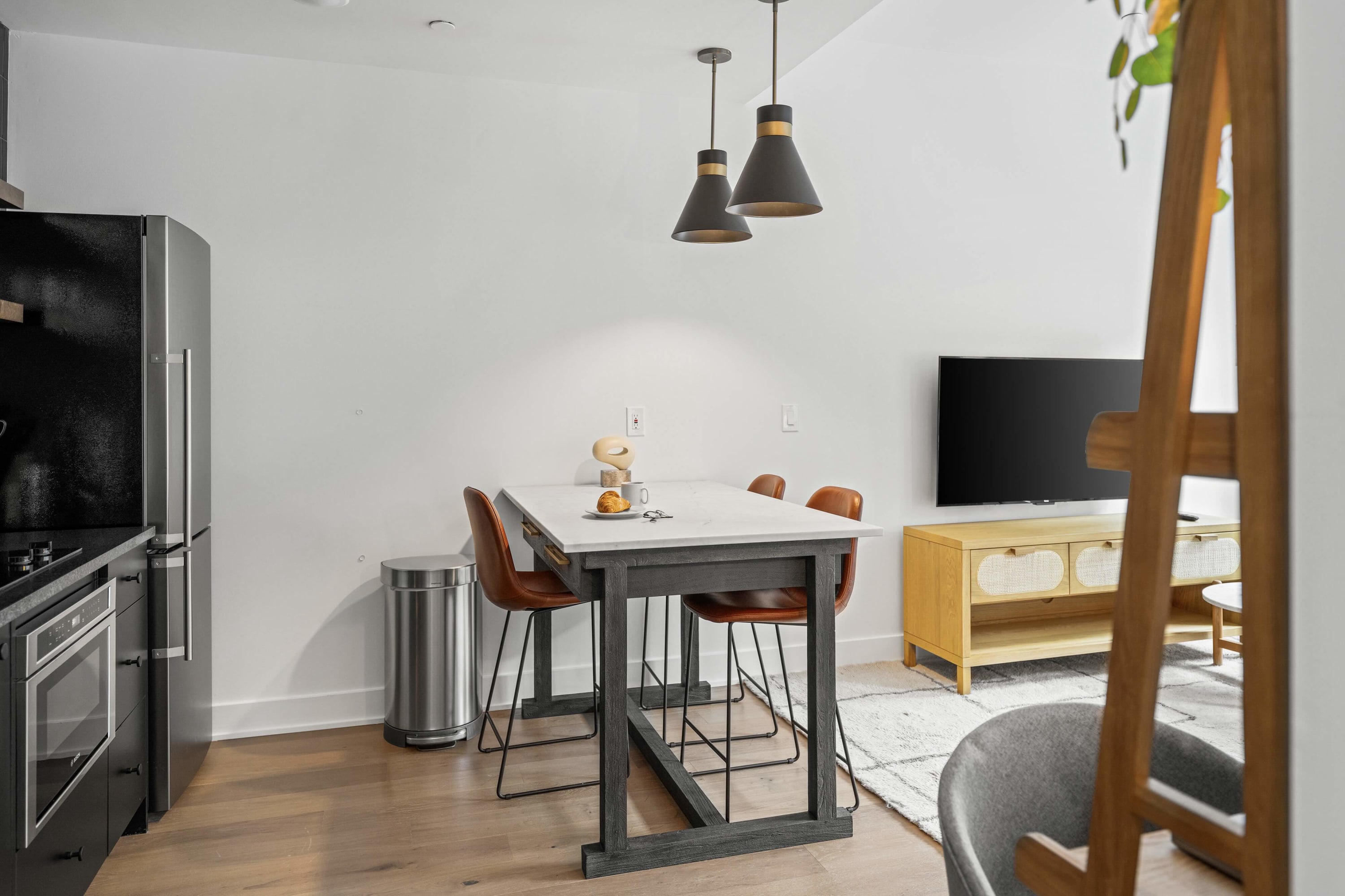 Open concept view of the modern kitchen and dining area, flowing into the living room. Features black cabinetry, stainless steel appliances, a dining island with orange stools, and a glimpse of the living room sofa and TV.