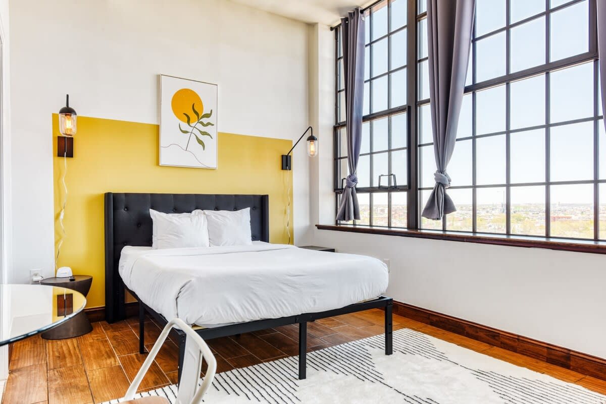 Bright studio bedroom featuring a queen bed with a dark upholstered headboard, a yellow accent wall with abstract art, and expansive industrial windows. Hardwood floors and a geometric area rug complete the modern and airy space.