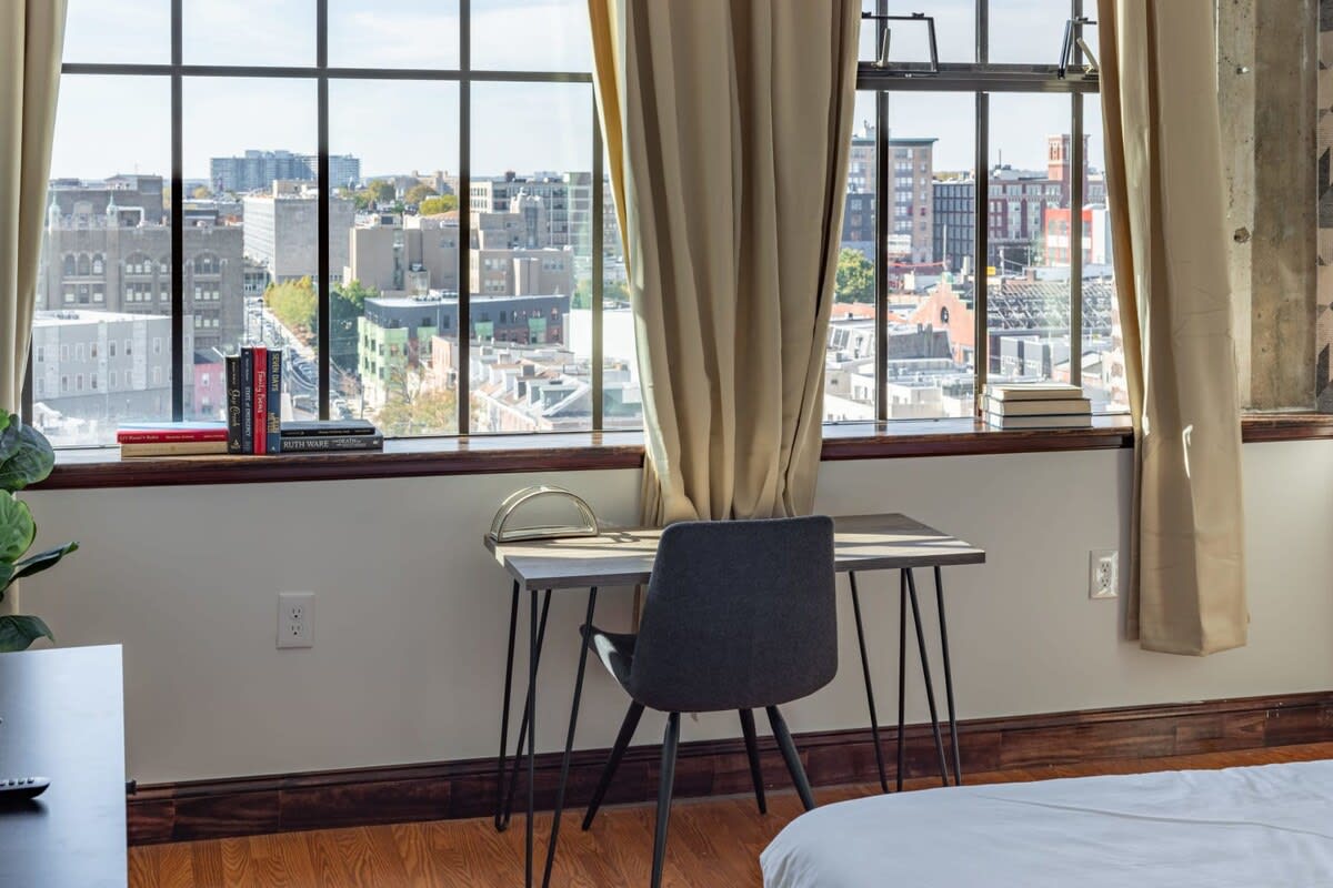 A close-up of a small, modern desk and chair positioned by a large industrial window, offering a panoramic view of the city skyline. Books are neatly stacked on the windowsill.