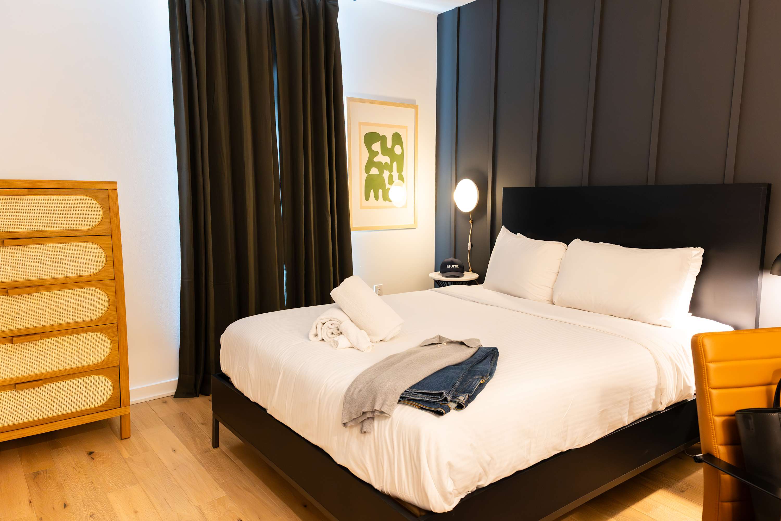 A cozy bedroom with a black bed frame, crisp white bedding, and a dark paneled accent wall behind the headboard. A light wood dresser with rattan drawer fronts stands on the left, while dark curtains frame the window.