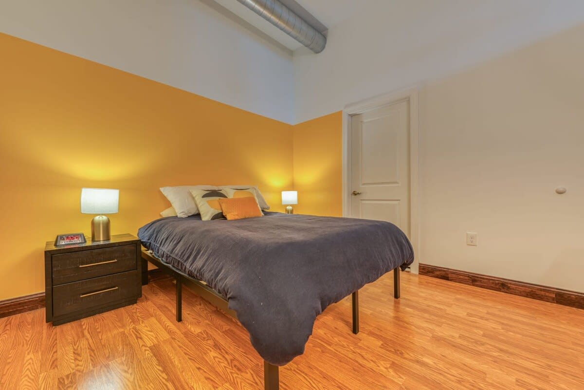 A bedroom with a dark blue bed frame and bedding is positioned against a vibrant yellow accent wall, contrasting with the white walls. Two nightstands with lamps flank the bed, and the room features light wood flooring.