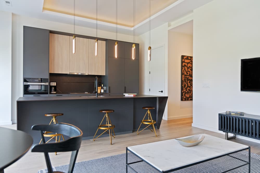 Open-concept living space showcasing a modern kitchen island with brass bar stools, a dark green sofa, a white marble coffee table, and a wall-mounted TV, with a large abstract artwork visible in the hallway beyond.