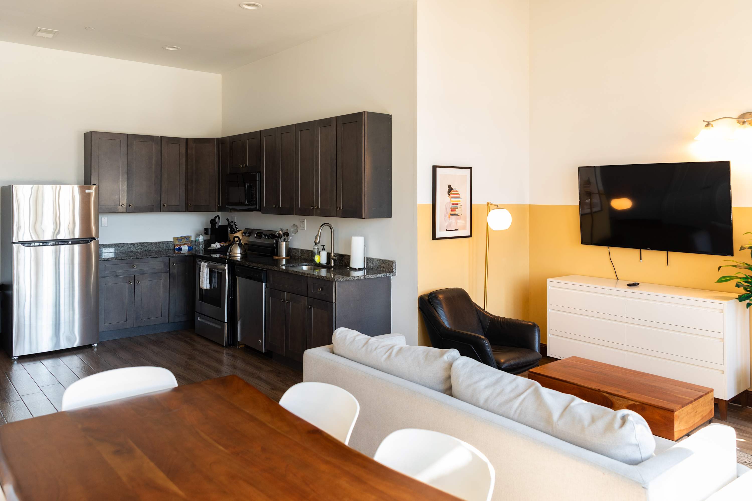 An open-concept living space with a modern kitchen, dining area, and living room with a light sofa and TV. A yellow accent wall adds a pop of color, complementing the dark cabinets and stainless steel appliances on hardwood floors.