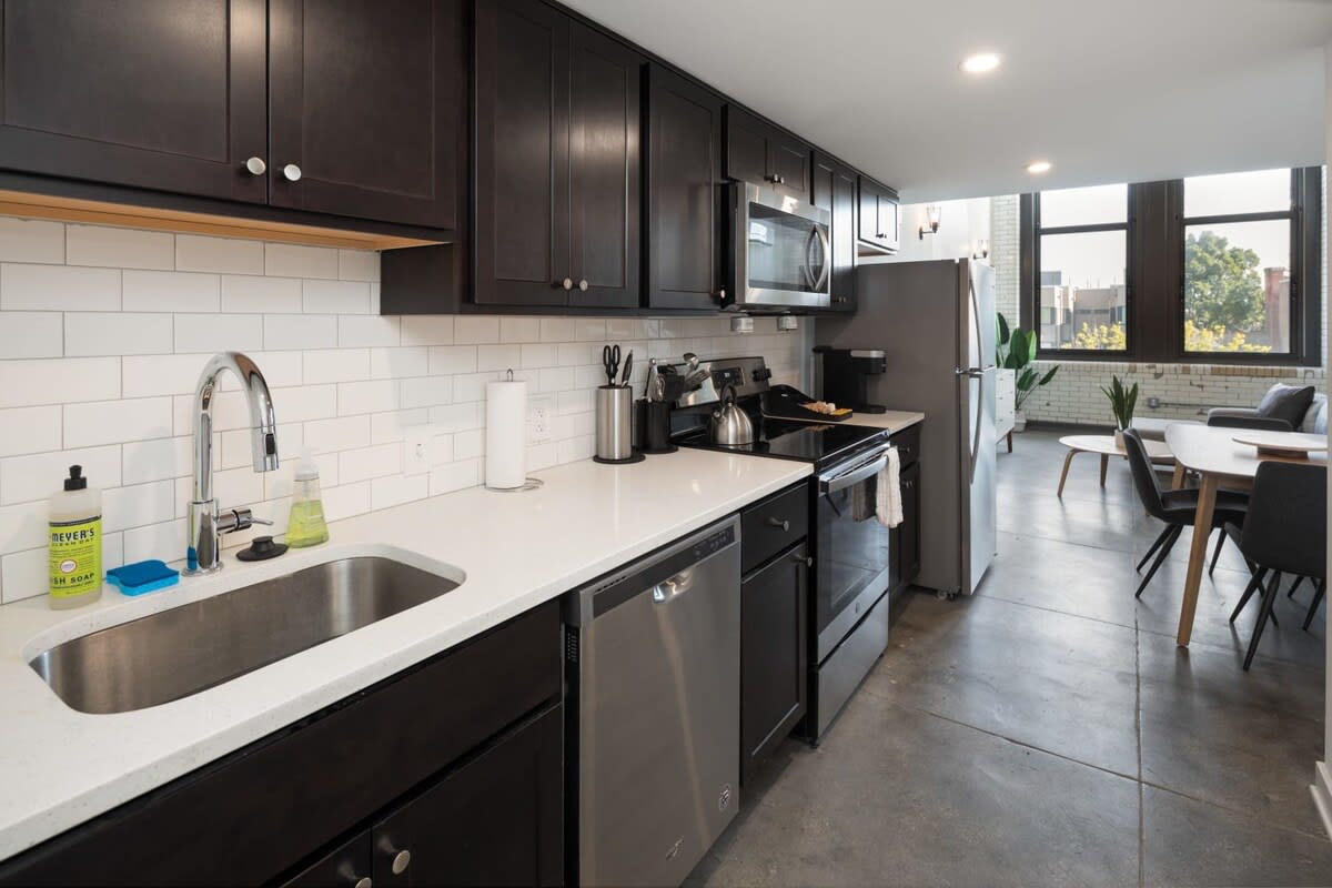 A wide shot of the modern kitchen with dark wood cabinets, white subway tile backsplash, and light quartz countertops, extending into the living area. Stainless steel appliances are visible, and concrete flooring runs throughout.