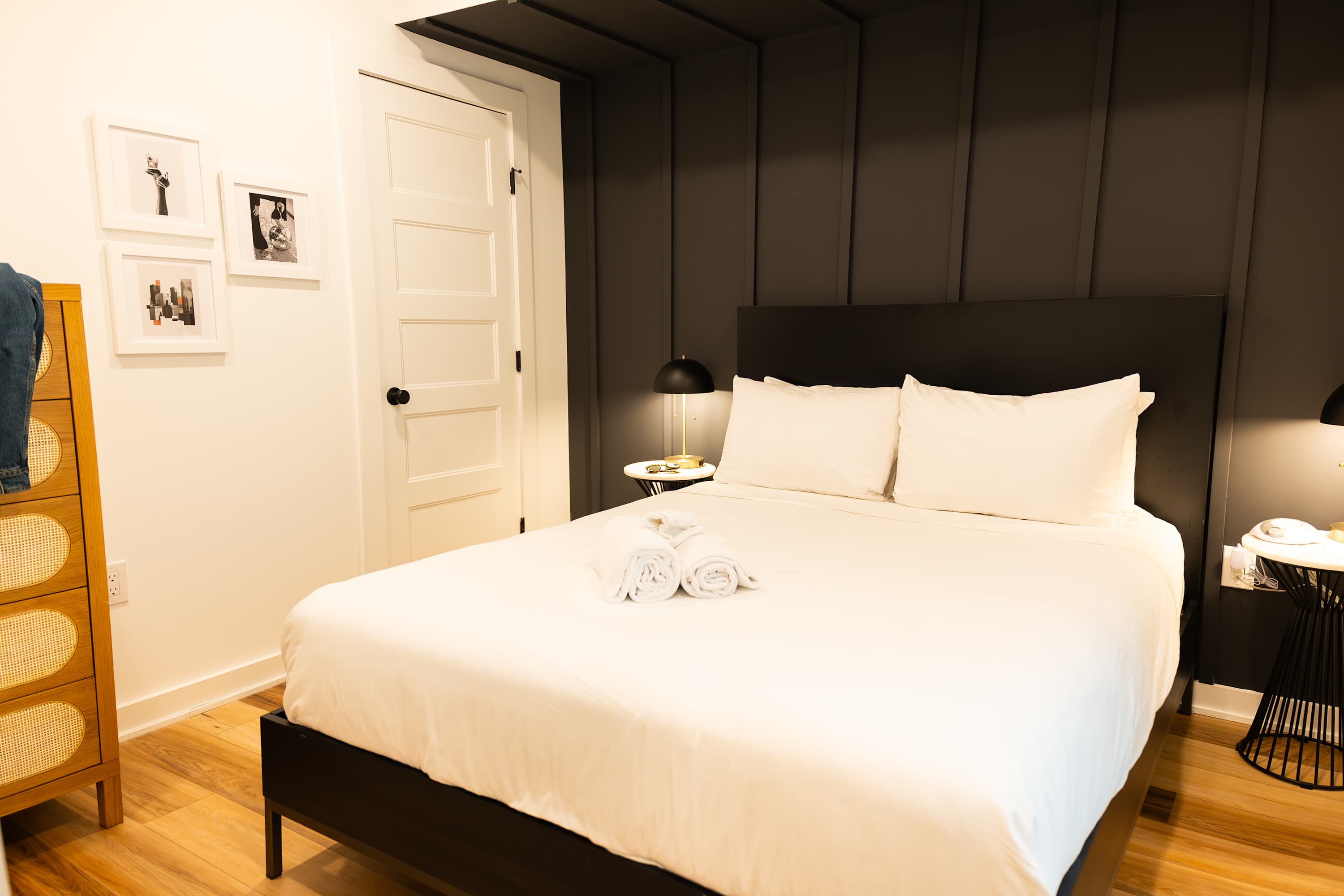 A modern bedroom featuring a queen-sized bed with white linens, a black headboard, dark paneled accent wall, and a white door, flanked by two nightstands with lamps.