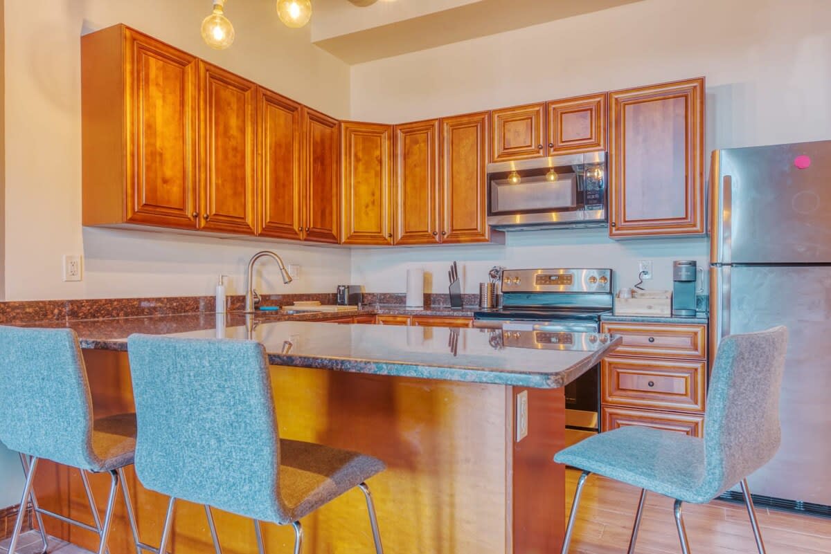 A well-equipped kitchen featuring warm-toned wooden cabinets, dark granite countertops, and stainless steel appliances including a microwave, range, and refrigerator. A sink with a gooseneck faucet is visible, and the kitchen island provides additional counter space.