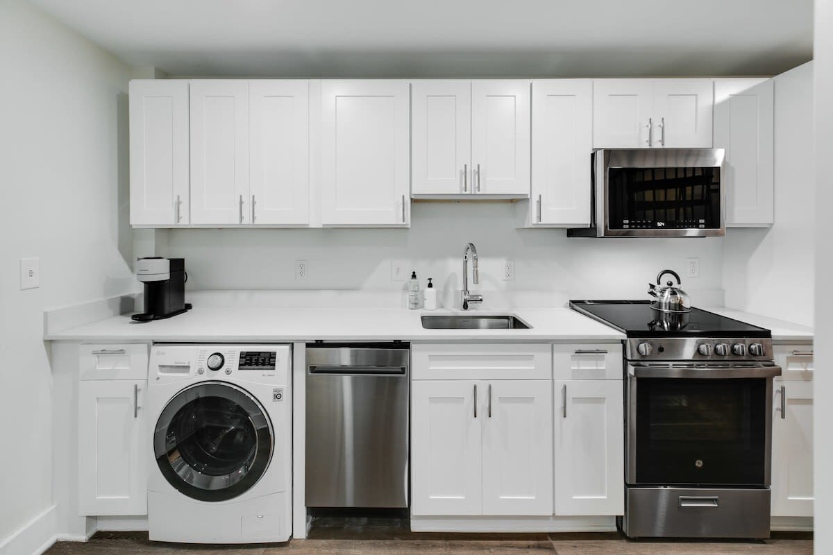A modern kitchen with white shaker cabinets, white countertops, and stainless steel appliances including a stove, microwave, dishwasher, and refrigerator. A front-loading washer and dryer are integrated into the counter space.