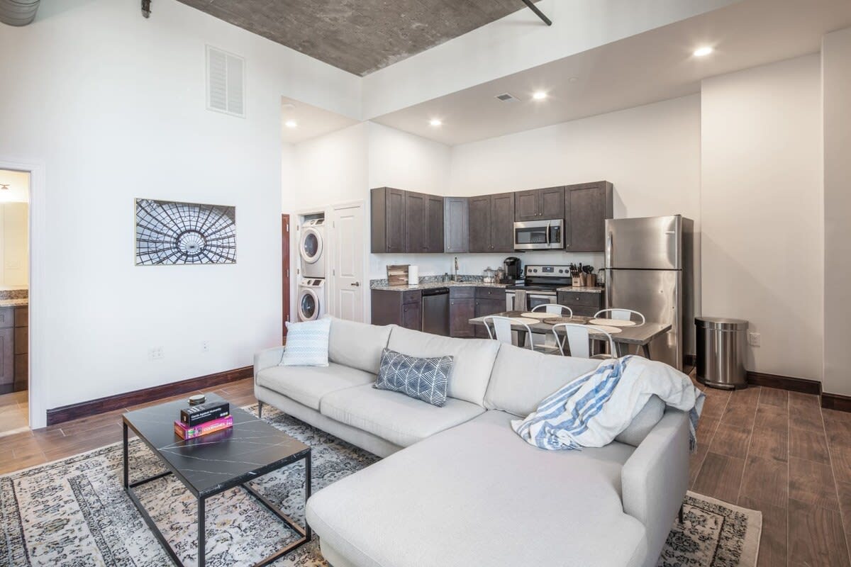 Bright open-plan living area with a light gray sectional sofa on an ornate rug, facing a black coffee table, with a view of the dark gray kitchen cabinets, stainless steel appliances, and a stacked washer and dryer unit in a nearby closet.