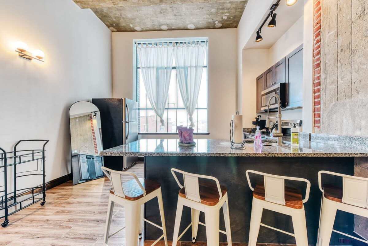 Kitchen area with a dark granite island and four white bar stools, positioned in front of a large window with sheer curtains. The kitchen features dark wood cabinets, stainless steel appliances, and wood-look flooring.