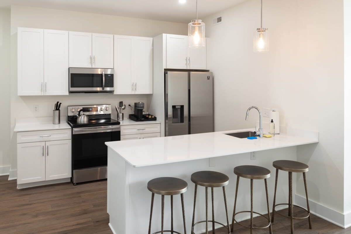 Modern kitchen with white shaker cabinets, stainless steel appliances, white quartz countertops, and a breakfast bar with three industrial-style bar stools.