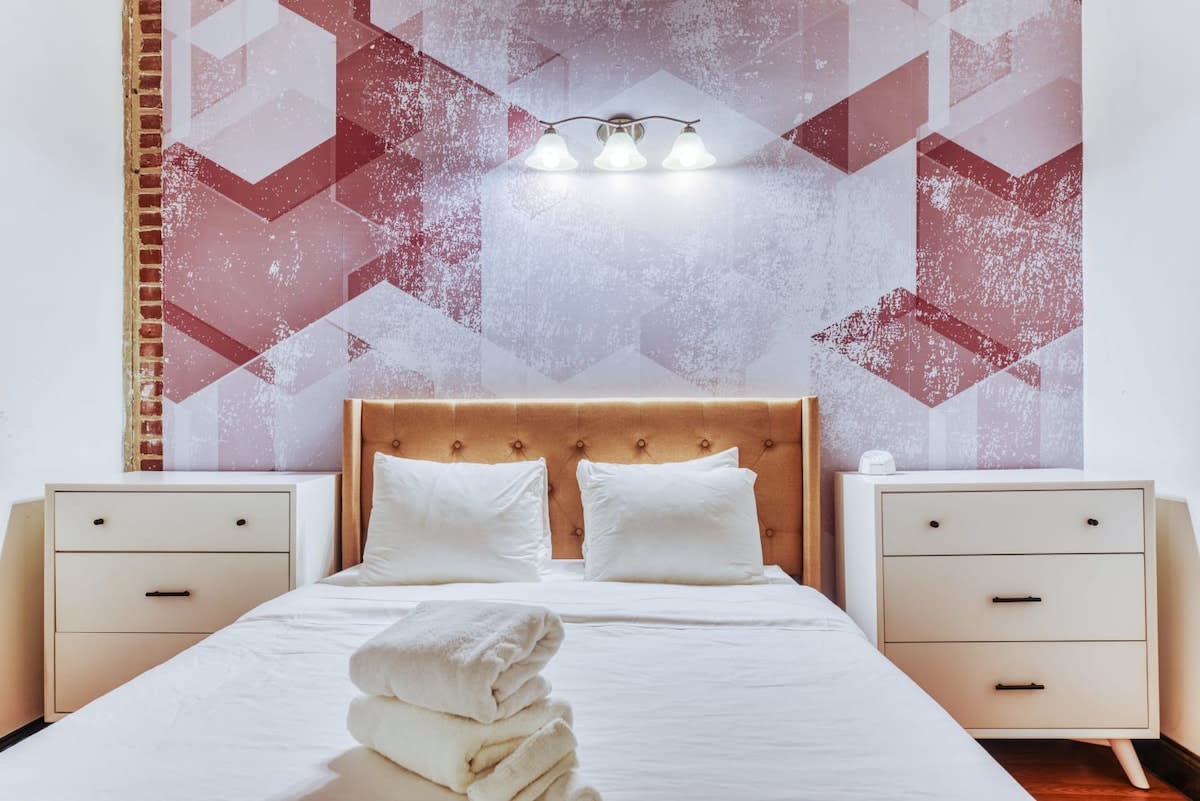 Bedroom with a queen bed featuring a tufted upholstered headboard and white bedding, accompanied by two white dressers serving as nightstands. A red and white geometric wallpaper covers the accent wall behind the bed.