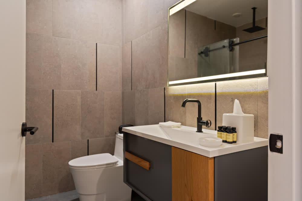 Modern bathroom featuring a white toilet, a vanity with a white countertop and dark cabinetry, and a rectangular mirror with integrated lighting. The walls are clad in large beige tiles with vertical black accents, complemented by black fixtures.