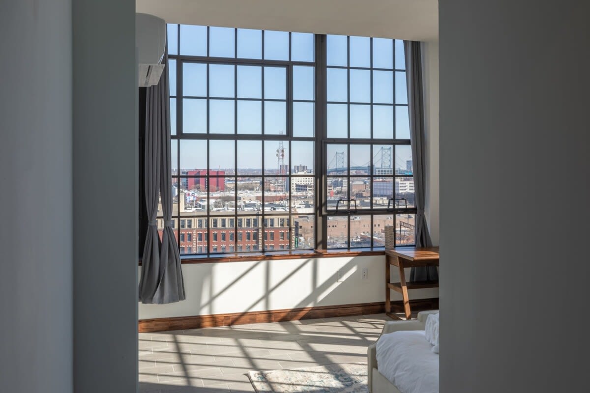 Interior view of the bright studio looking towards expansive industrial-style grid windows that frame a wide city panorama, including a distant bridge. Sunlight casts long shadows across the light-colored tiled floor.