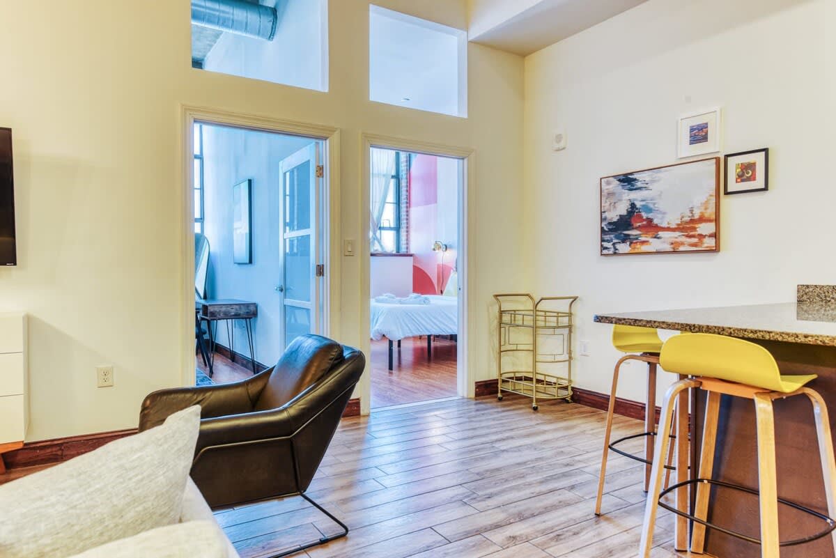 A wide view of the open-concept living area, showing a light grey sofa and a dark leather armchair on a patterned rug, with a glimpse of the kitchen's breakfast bar and yellow stools. Two doorways with glass panels lead into separate bedrooms, one with a colorful accent wall visible.