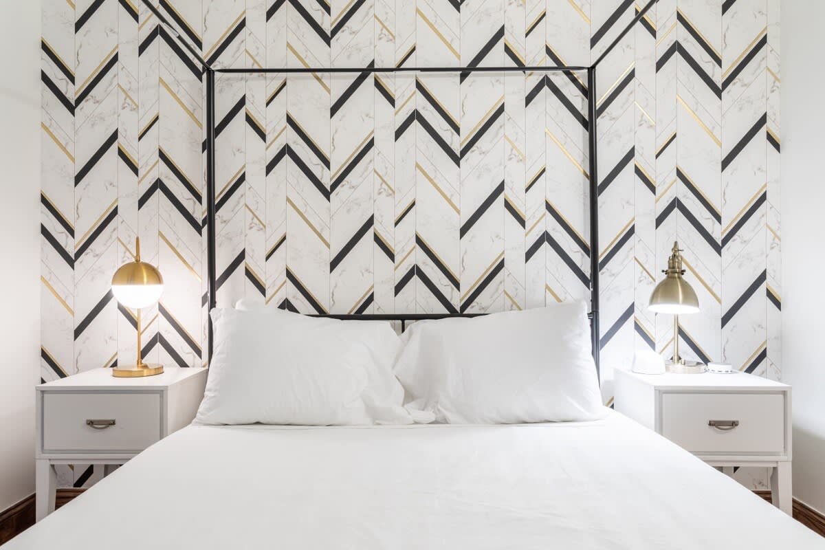 Modern bedroom featuring a bed with a black metal canopy frame, white bedding, two white nightstands with gold lamps, and a striking black, white, and gold chevron patterned wallpaper behind the bed.