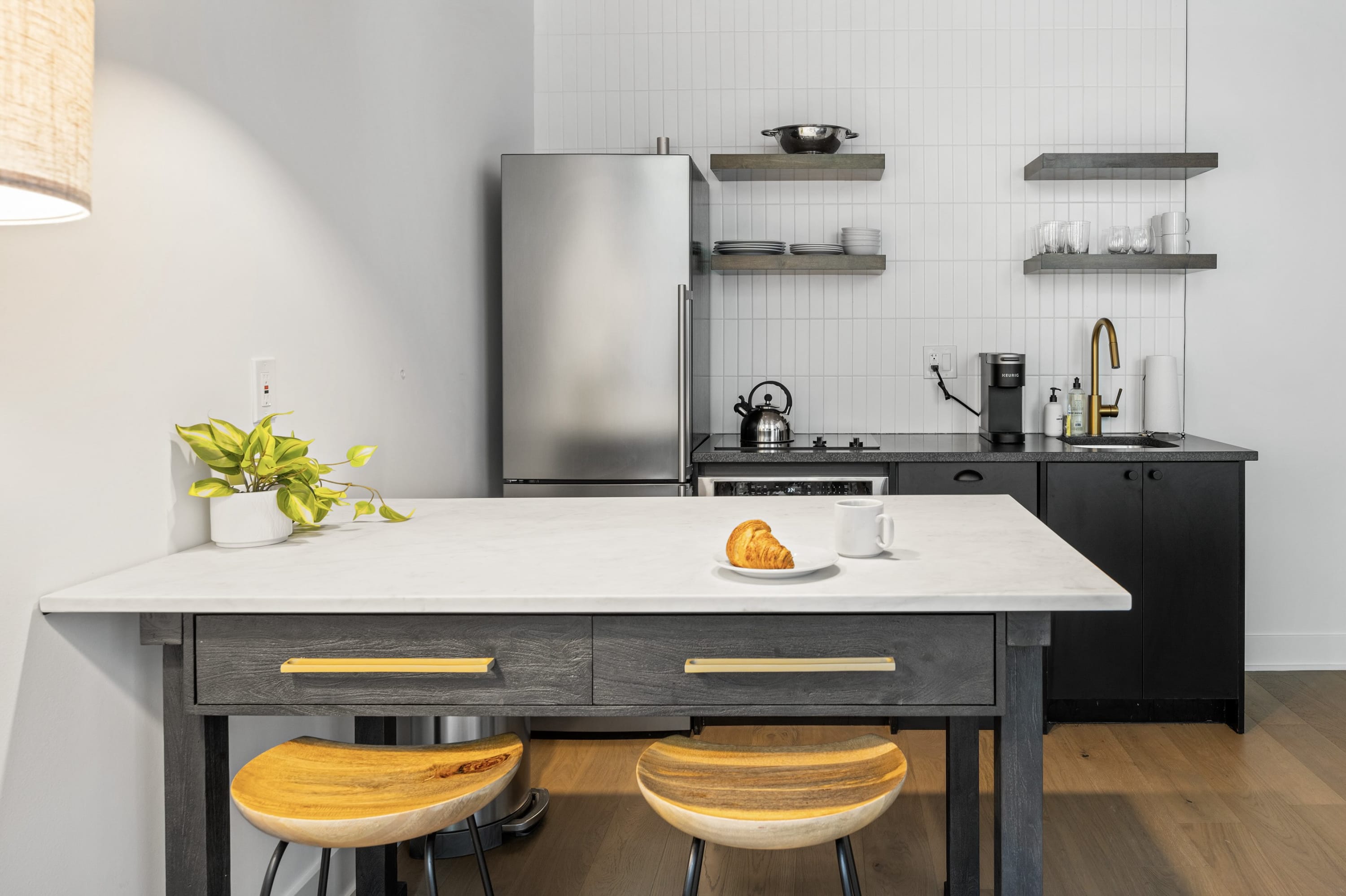 A compact, modern kitchen with black cabinetry, white subway tile backsplash, and stainless steel appliances including a refrigerator and built-in oven. Open wooden shelves display dishes, and a dining table with stools is in the foreground.
